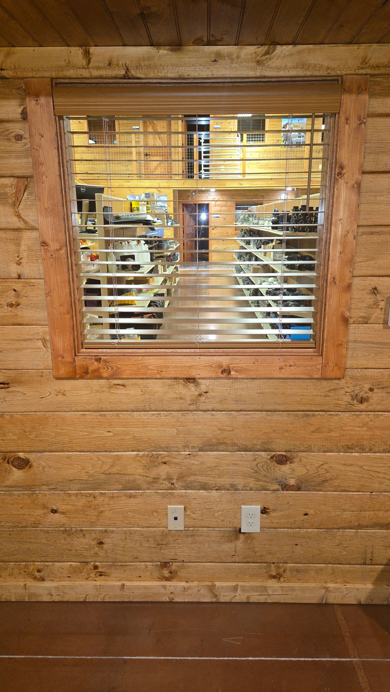 Rustic wooden cabin wall with a window overlooking a lit interior and staircase behind blinds.