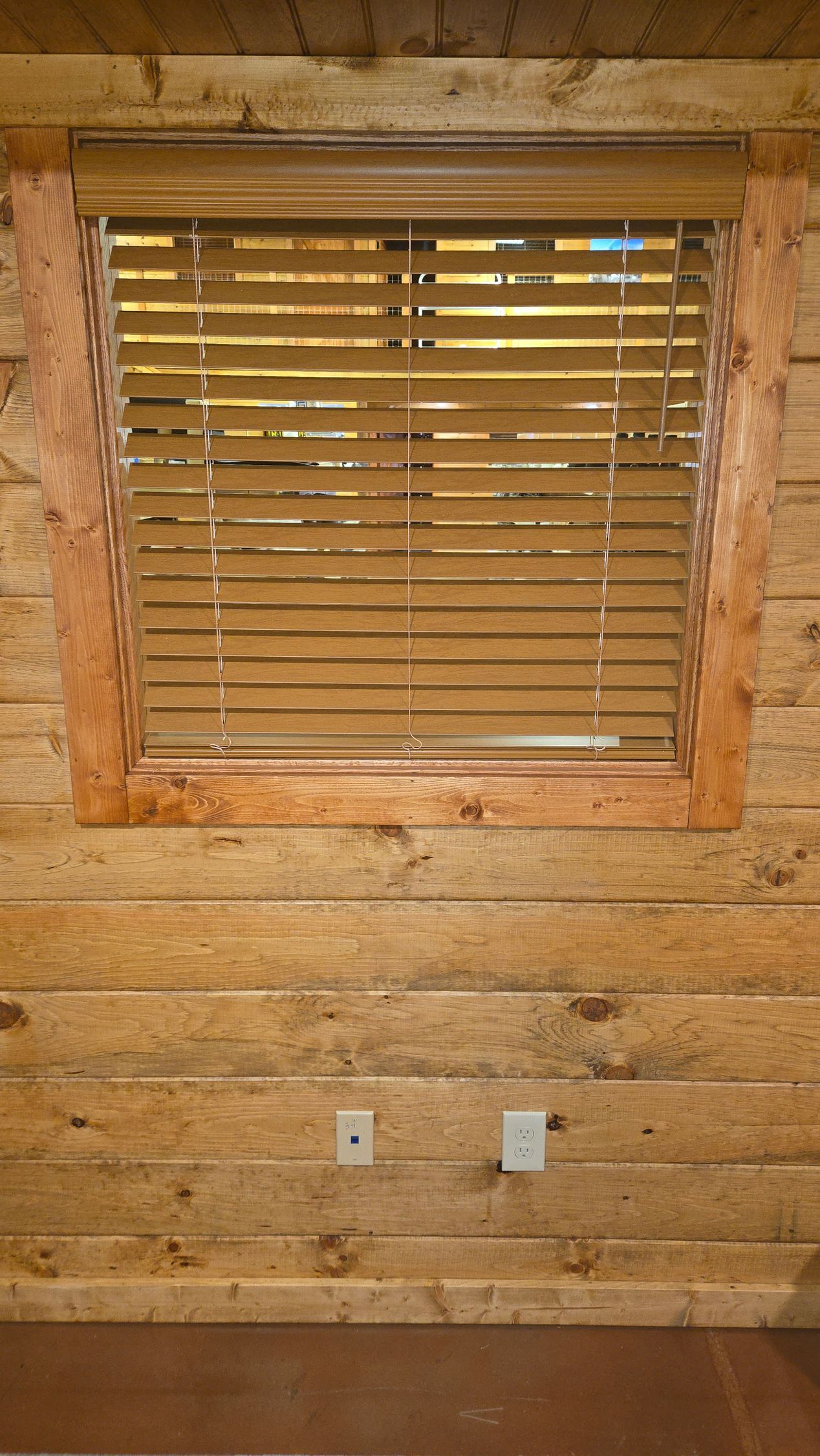 Wood-paneled wall with a small window covered by wooden blinds and two outlets below.