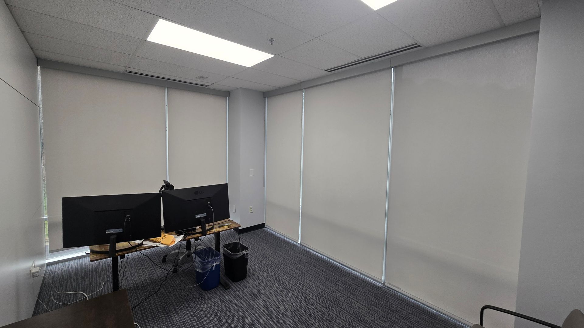 Office with two monitors on a desk, bags on the floor, and closed window blinds under bright ceiling lights