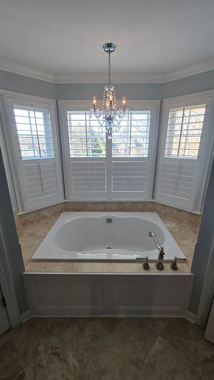 Bright bathroom with a white soaking tub, chandelier, bay windows, and light gray walls.