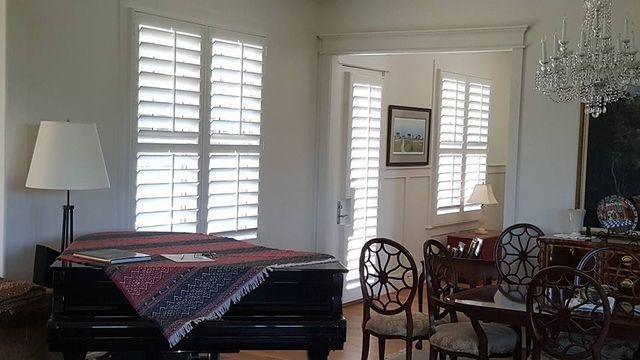 Sunlit dining room with piano, wooden chairs, table, lamp, and chandelier near shuttered windows