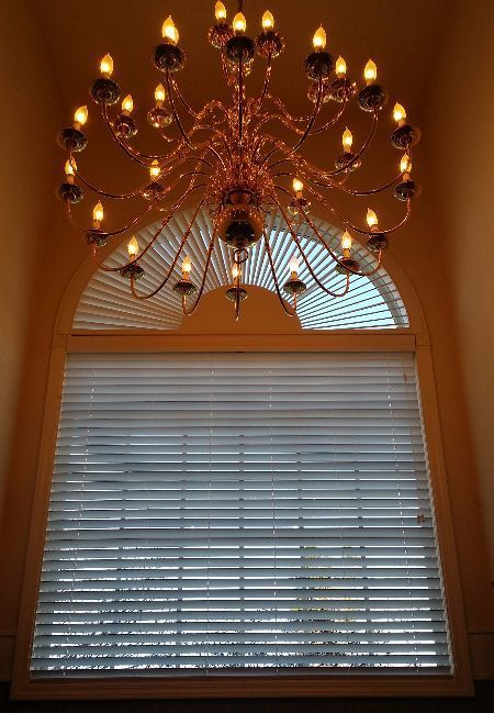 Chandelier hanging above a window with closed white blinds and an arched transom in a warm-toned room