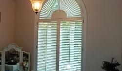 Interior wall with arched window, white shutters, and a lit wall sconce