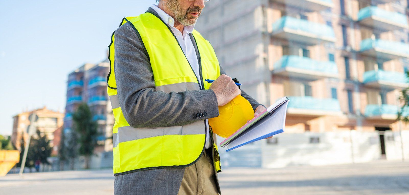 Construction worker in a yellow vest, writing on a clipboard, holding a hard hat in front of a building under construction.