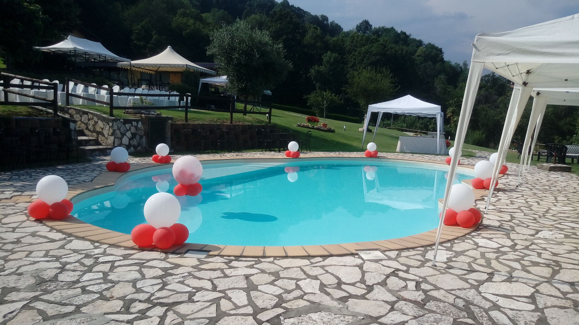 Piscina decorata con palloncini rossi e bianchi, circondata da un patio in pietra, gazebo e una verde collina.