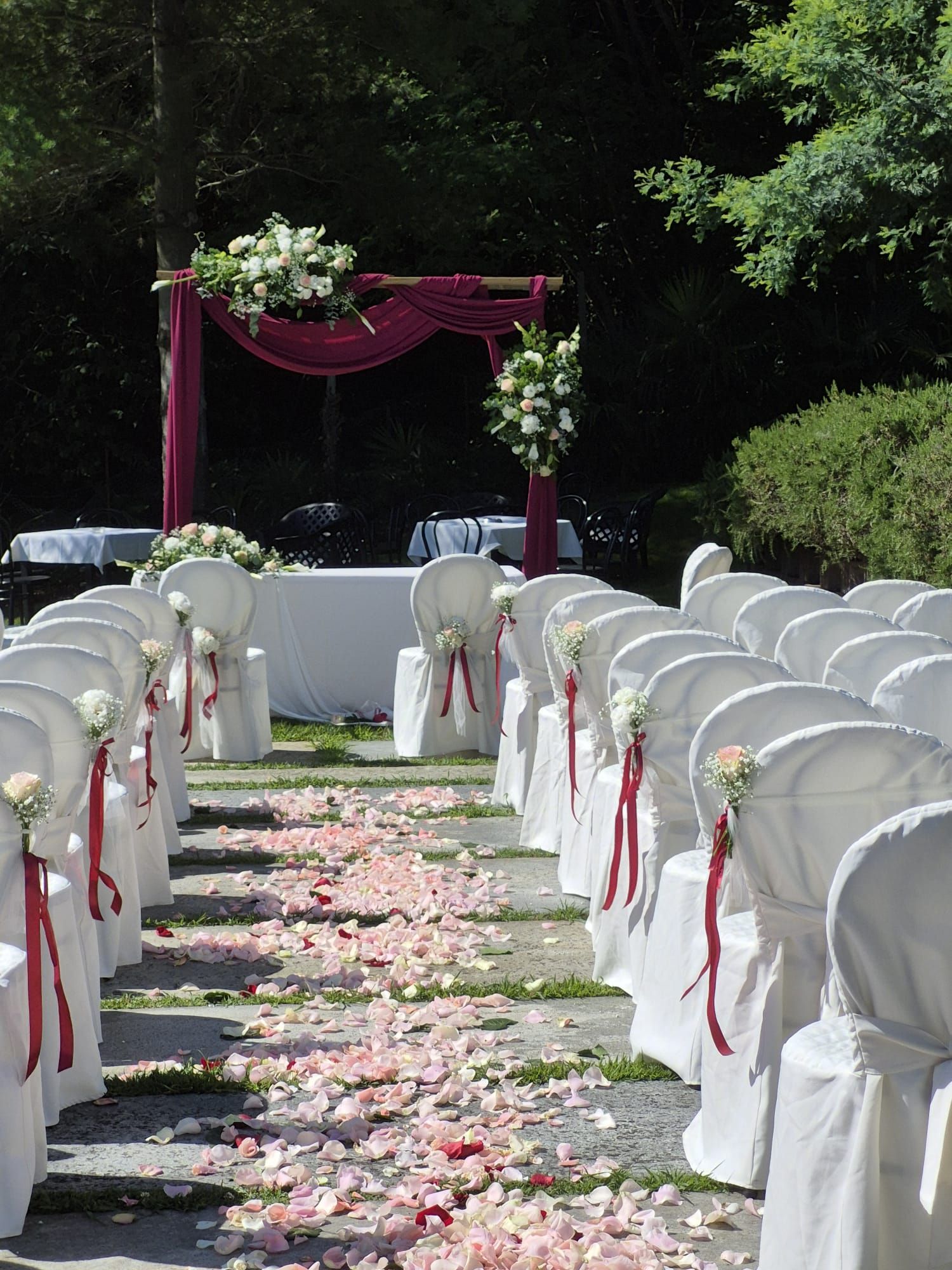 Allestimento per la cerimonia nuziale con sedie bianche, petali di rose rosa e una chuppah color bordeaux.