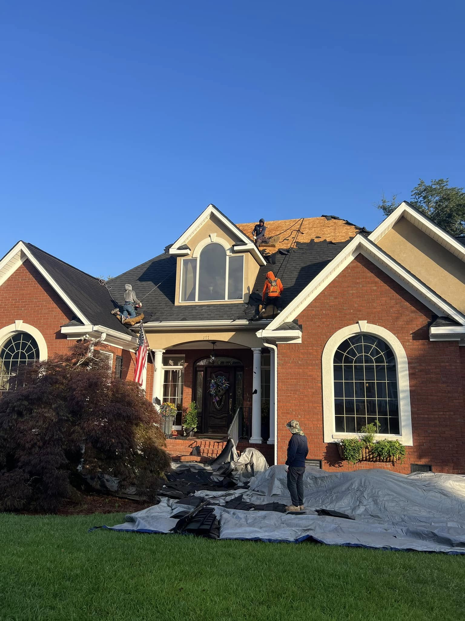 House with roof damage, workers on roof. Man standing on ground with debris and blue sky.