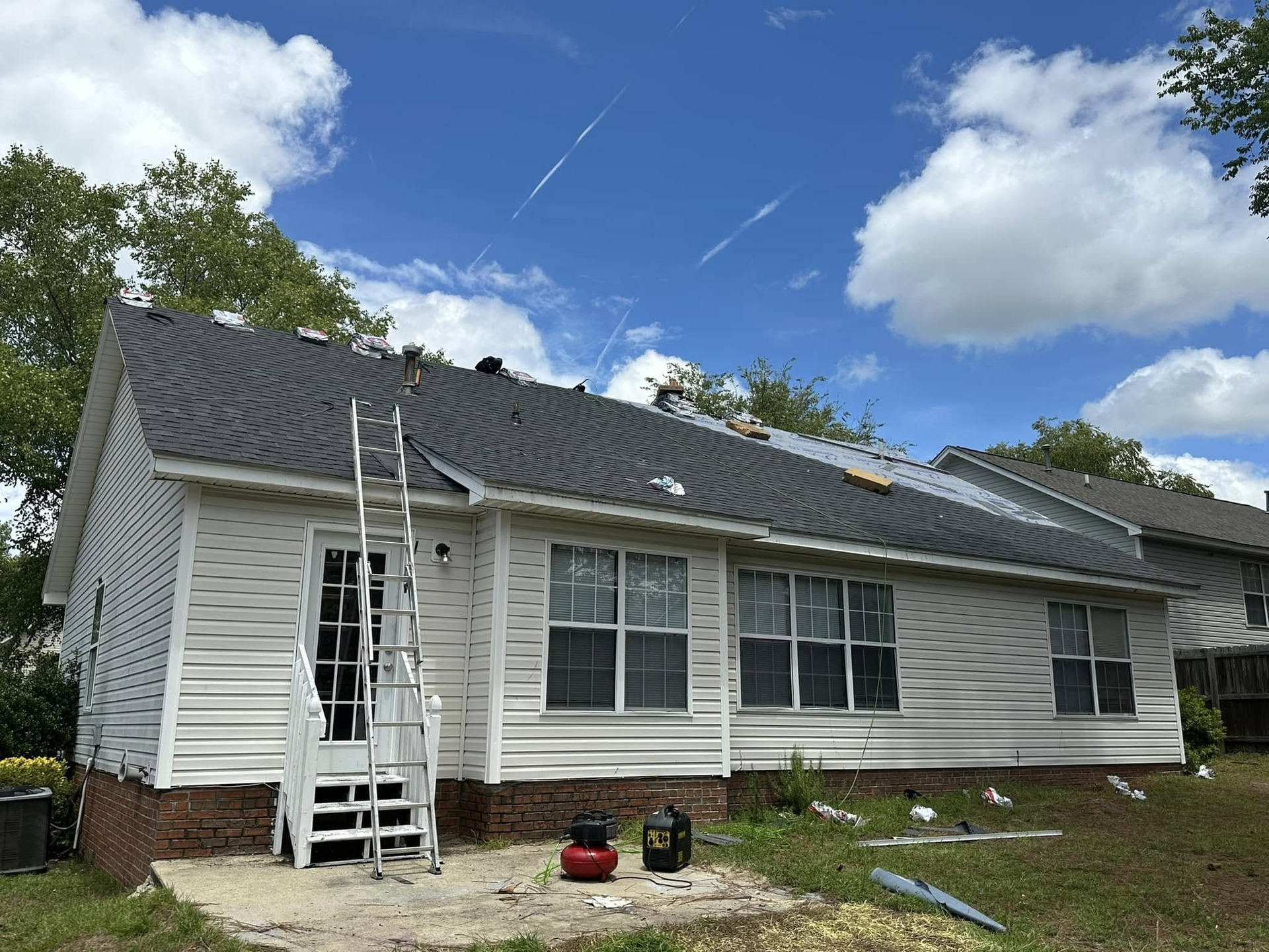 House with roof partially replaced, ladder leaning against door. Blue sky, white clouds.