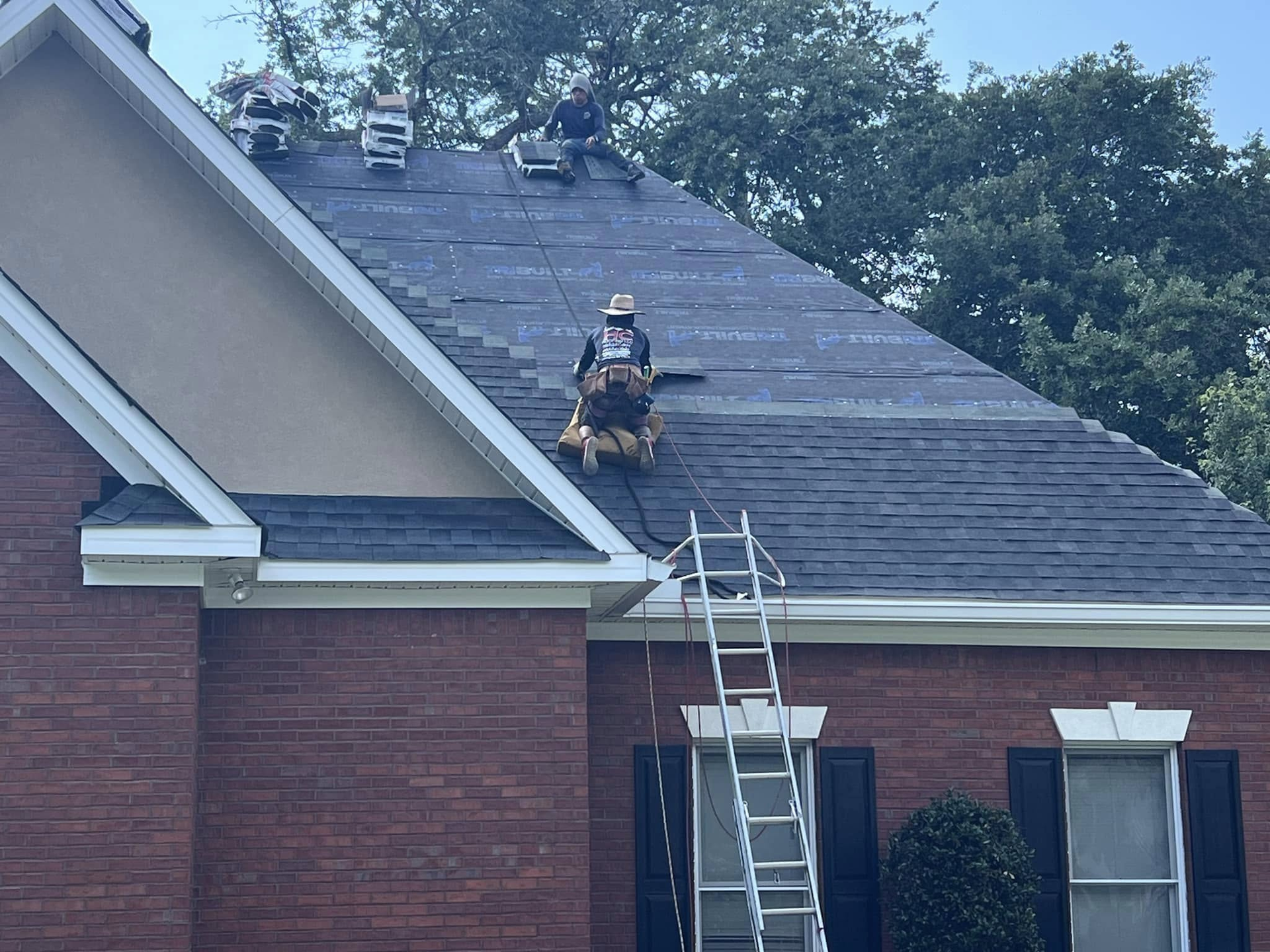 Firefighters on a roof with a ladder, working on a dark shingle roof with some missing shingles, in front of trees.