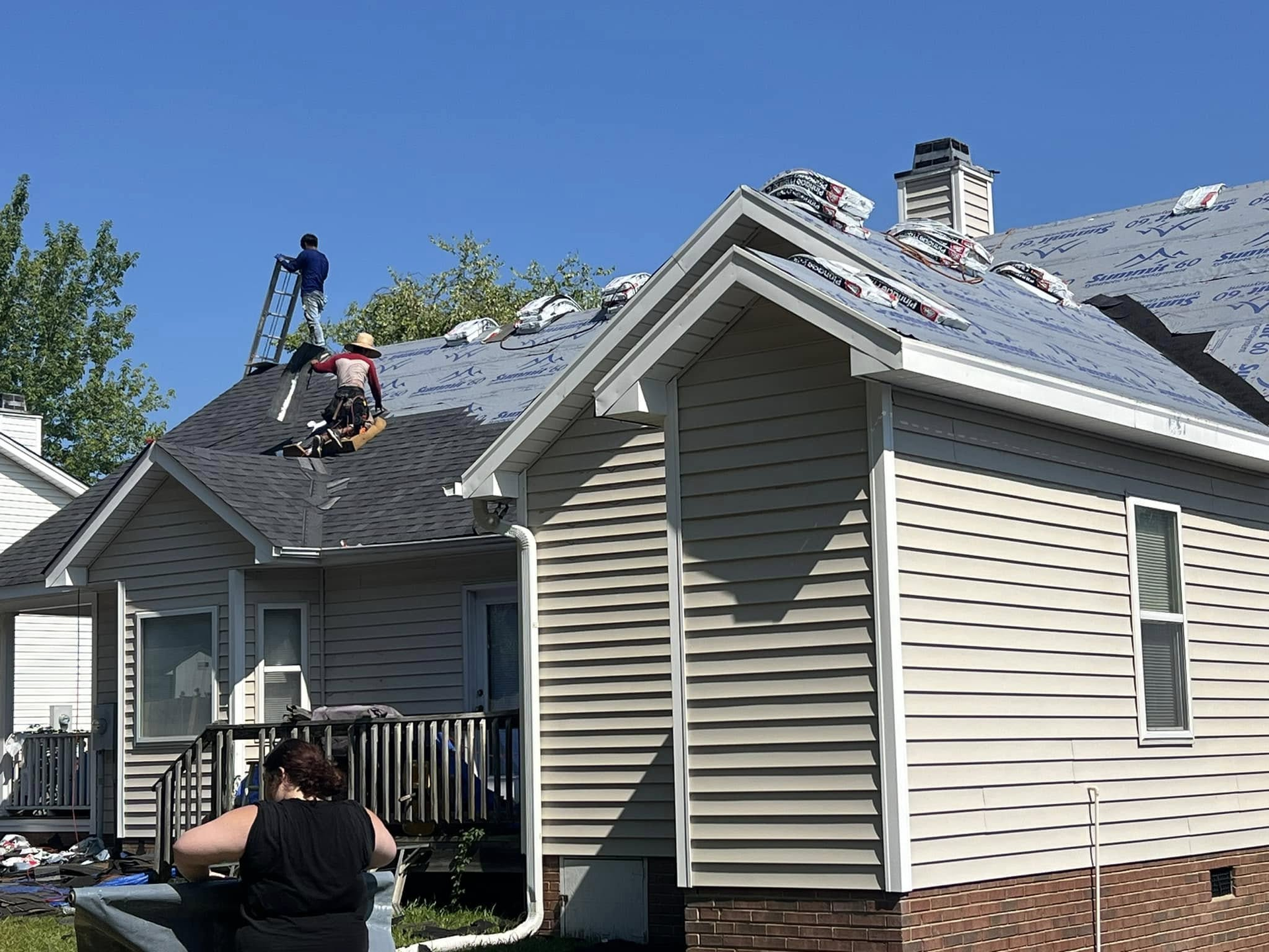 Workers replacing a roof on a house with gray siding under a clear blue sky.