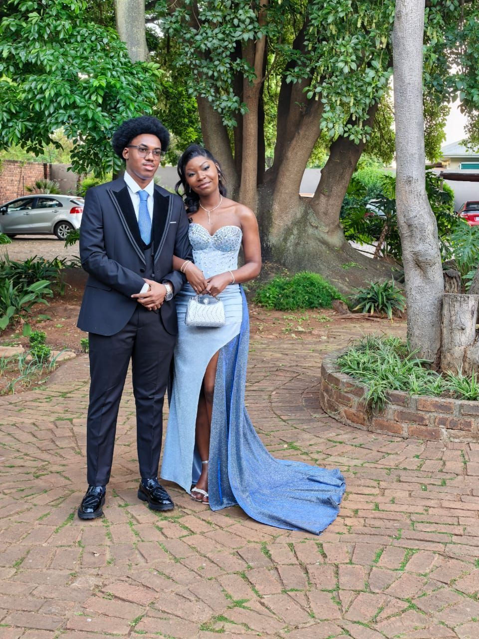 Two people pose in formal attire outdoors on a brick path beside trees and greenery.