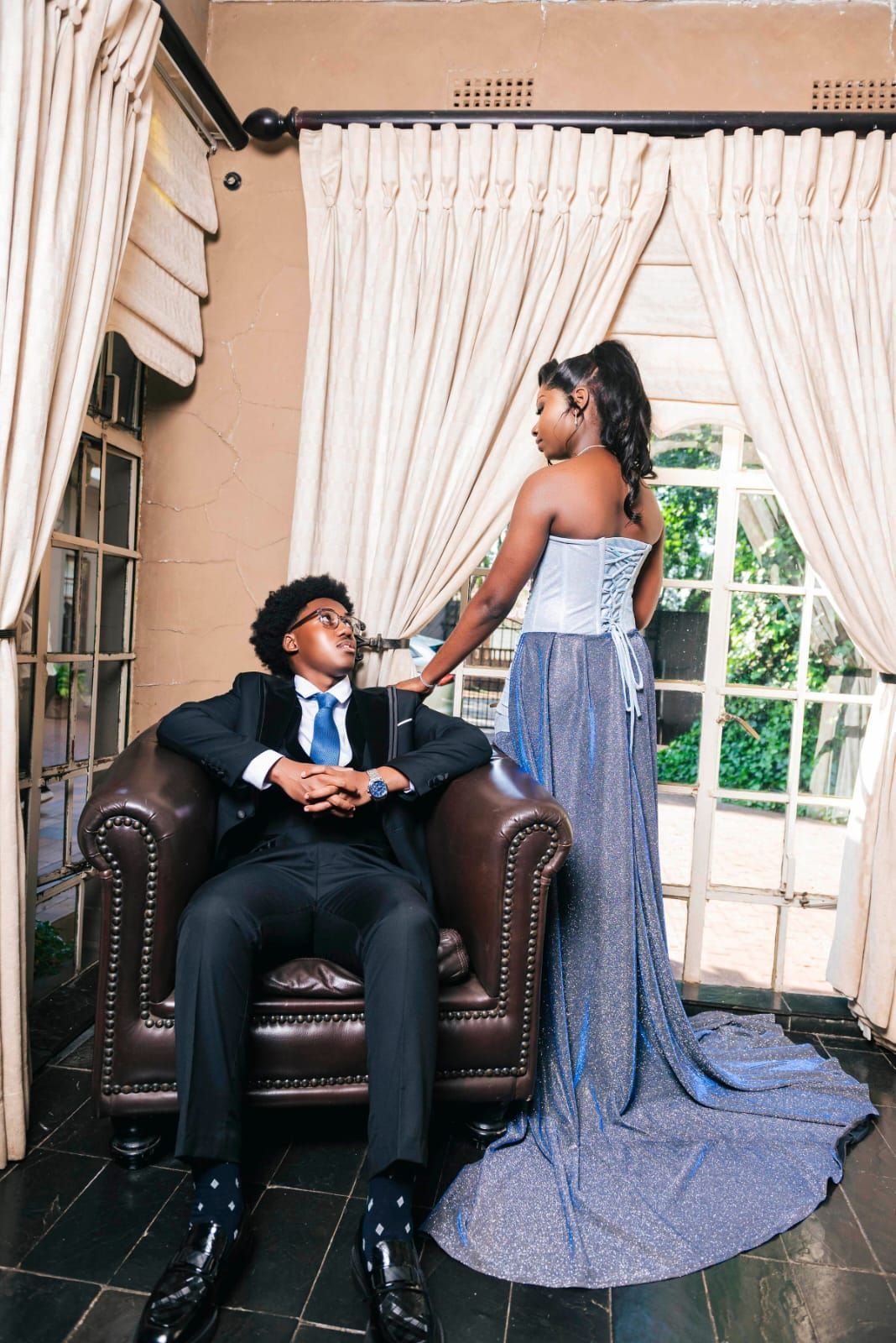 Two people posing indoors; one sits in a dark armchair while the other stands beside them in a sparkling blue gown.