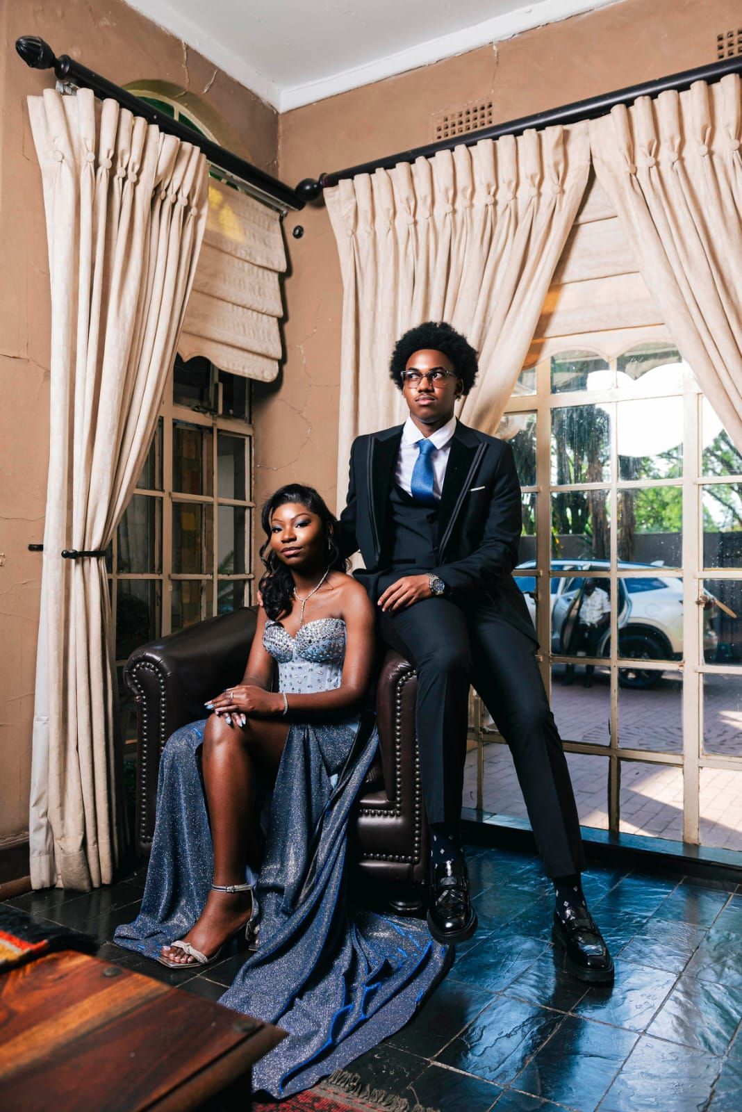 Two people in formal attire pose indoors by tall windows with cream curtains.