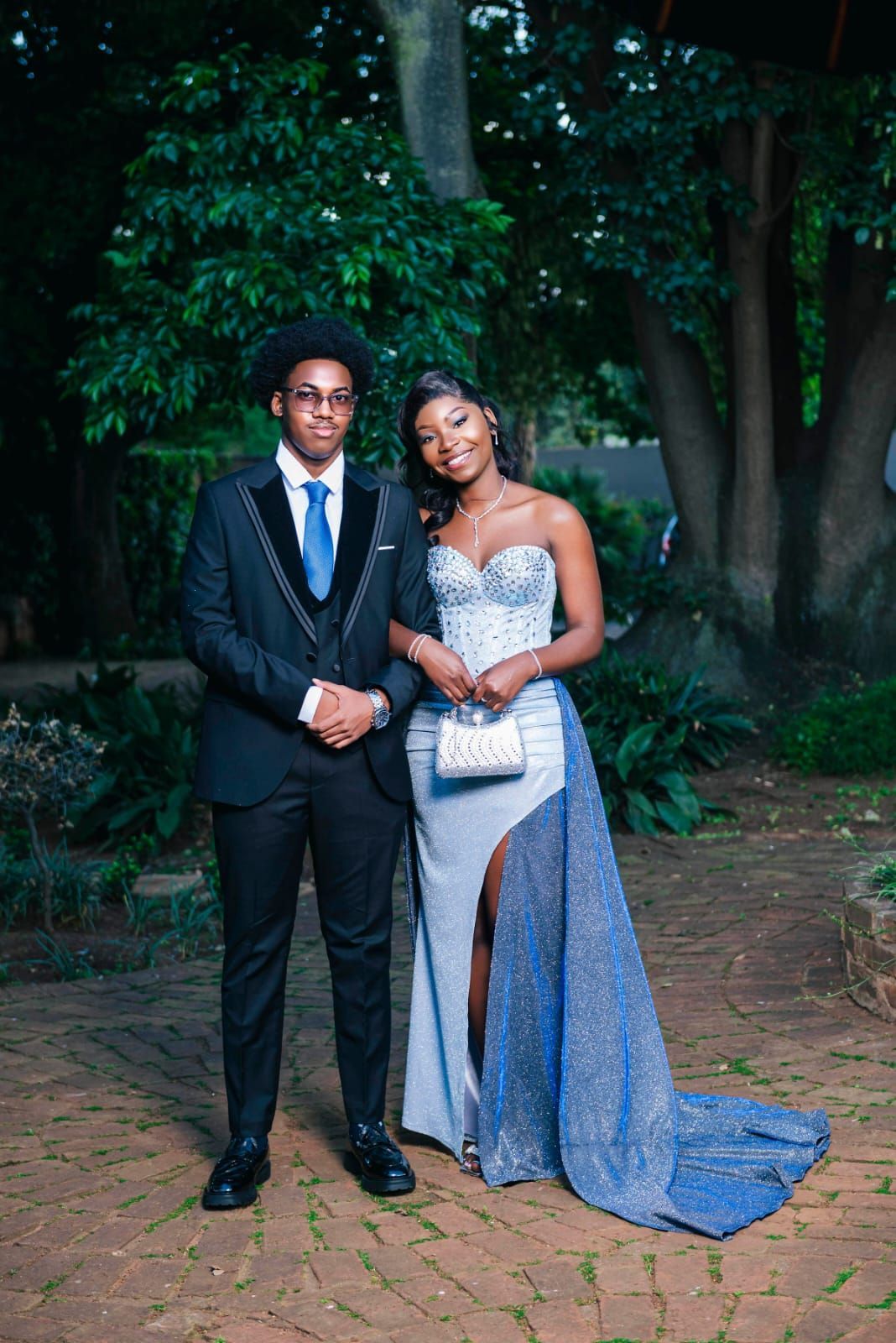 Couple posing outdoors in formal attire, with the woman in a blue gown holding a clutch.