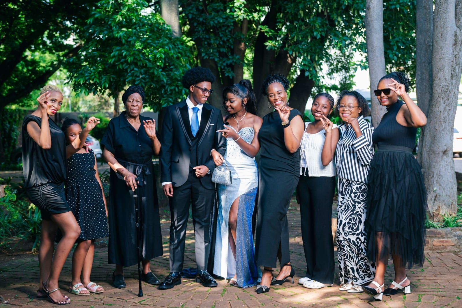 Group in formal black-and-white outfits posing and smiling outdoors under trees