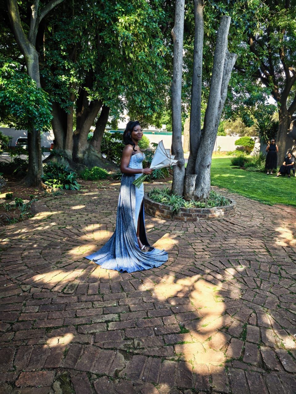 Woman in a blue gown holding a bouquet in a garden with trees and a brick path