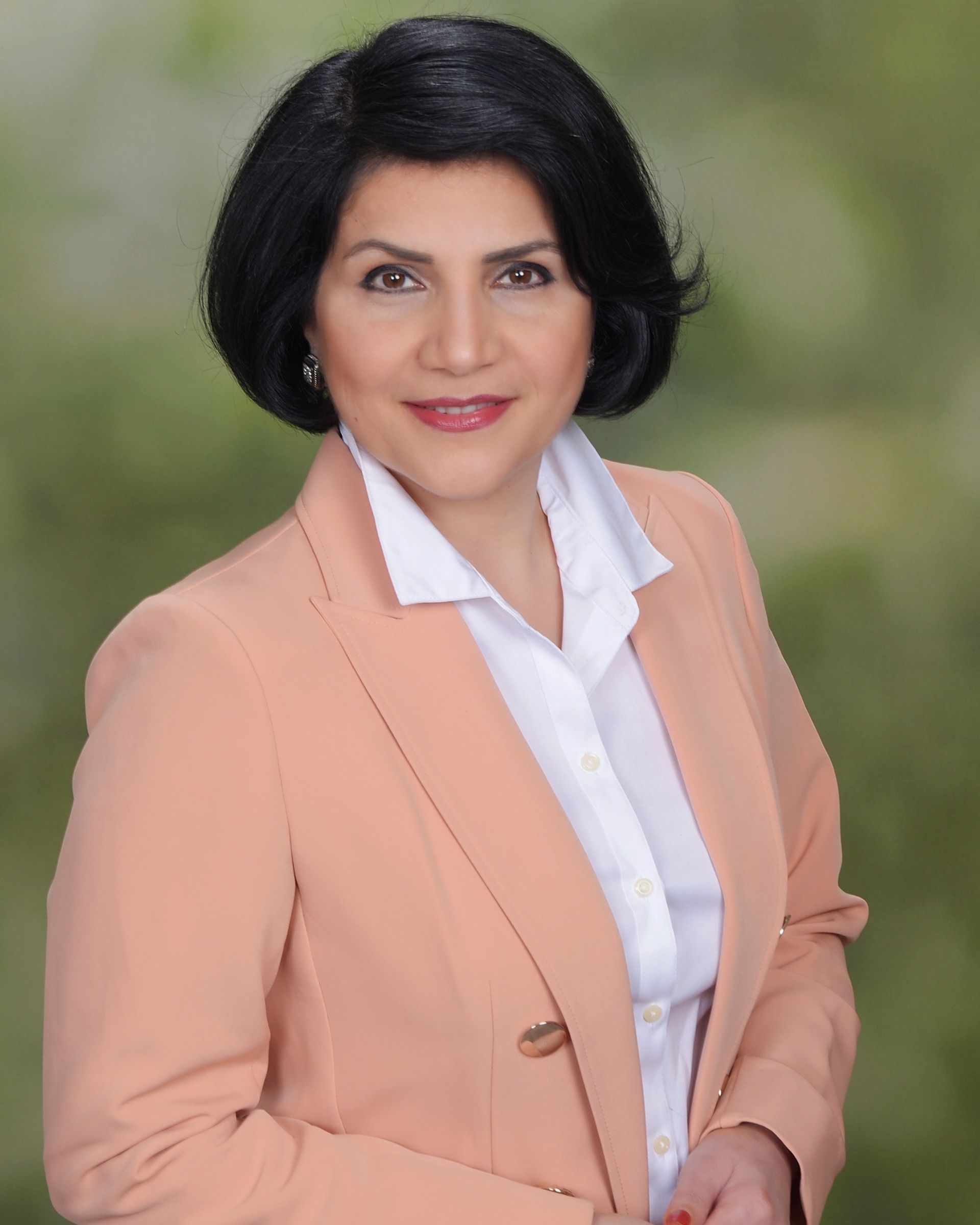 Woman in a peach blazer and white shirt, smiling, set against a blurred green backdrop.