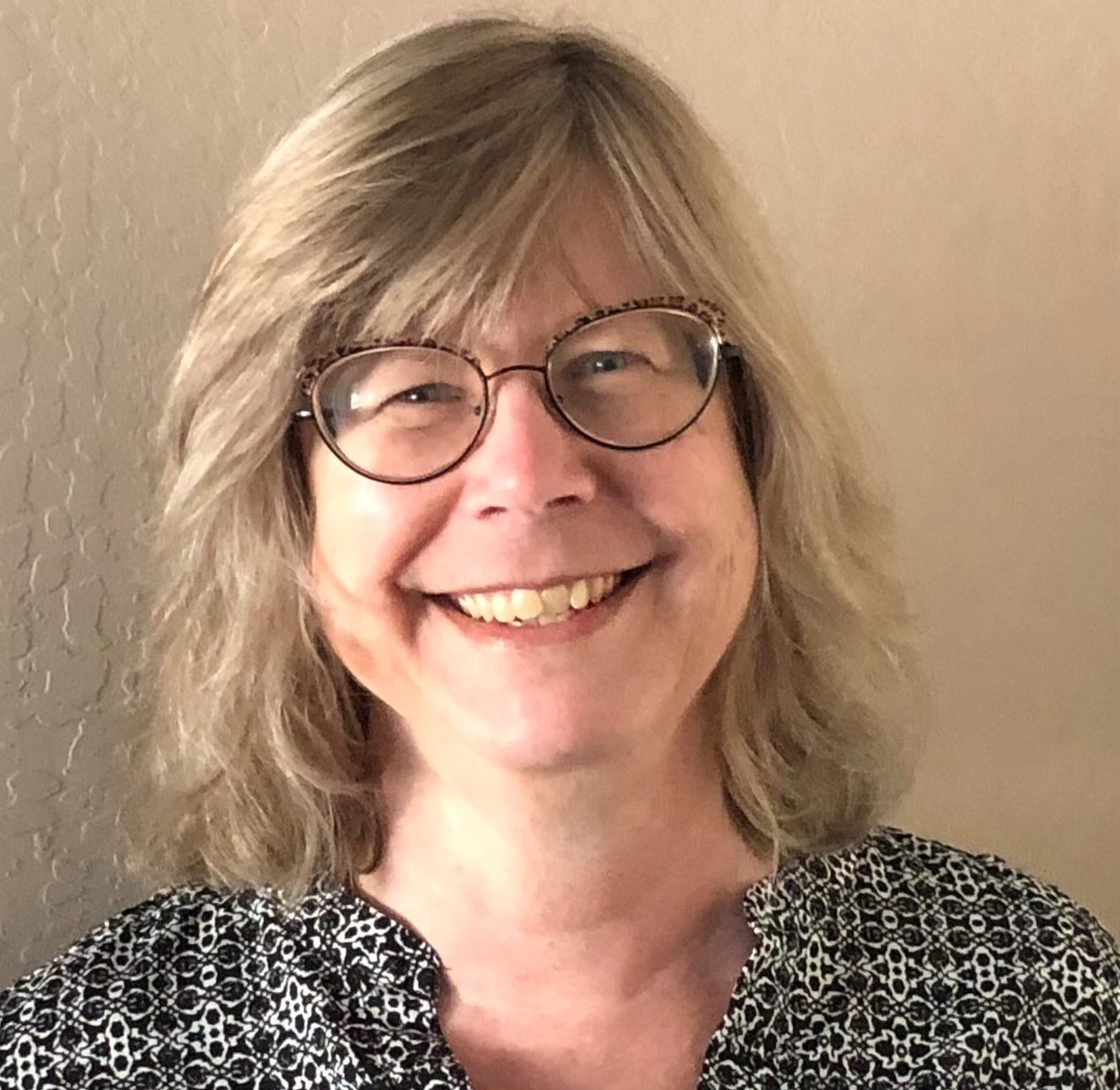 Woman with glasses smiles; blonde hair, patterned top, soft background.