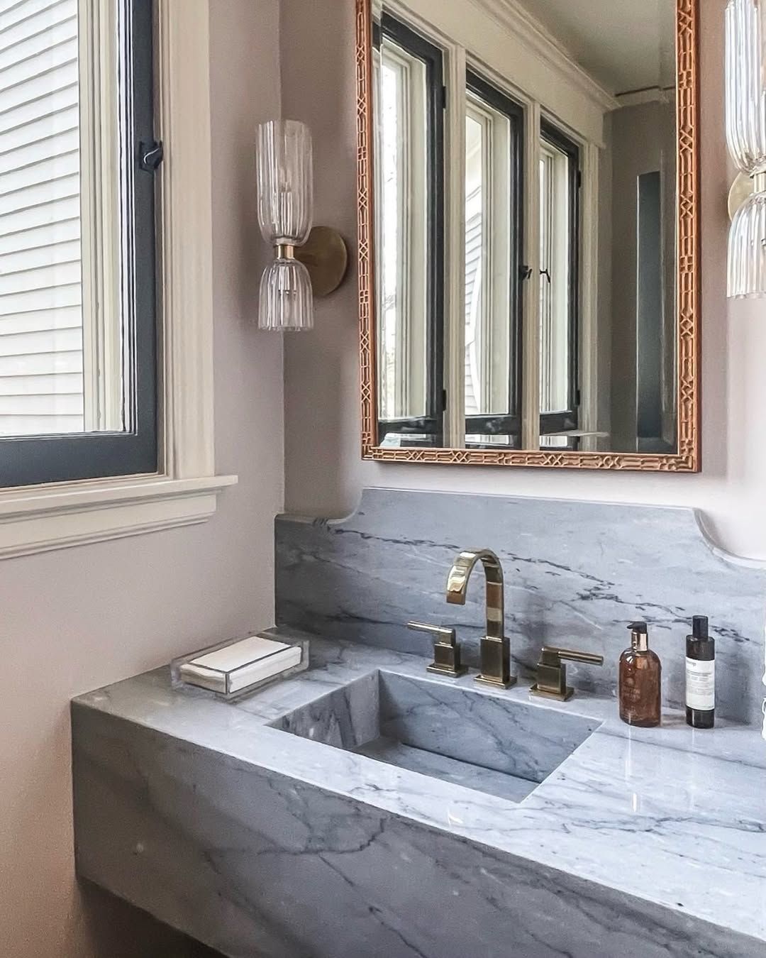 Bathroom with marble vanity, gold fixtures, mirror, window, and blush walls.
