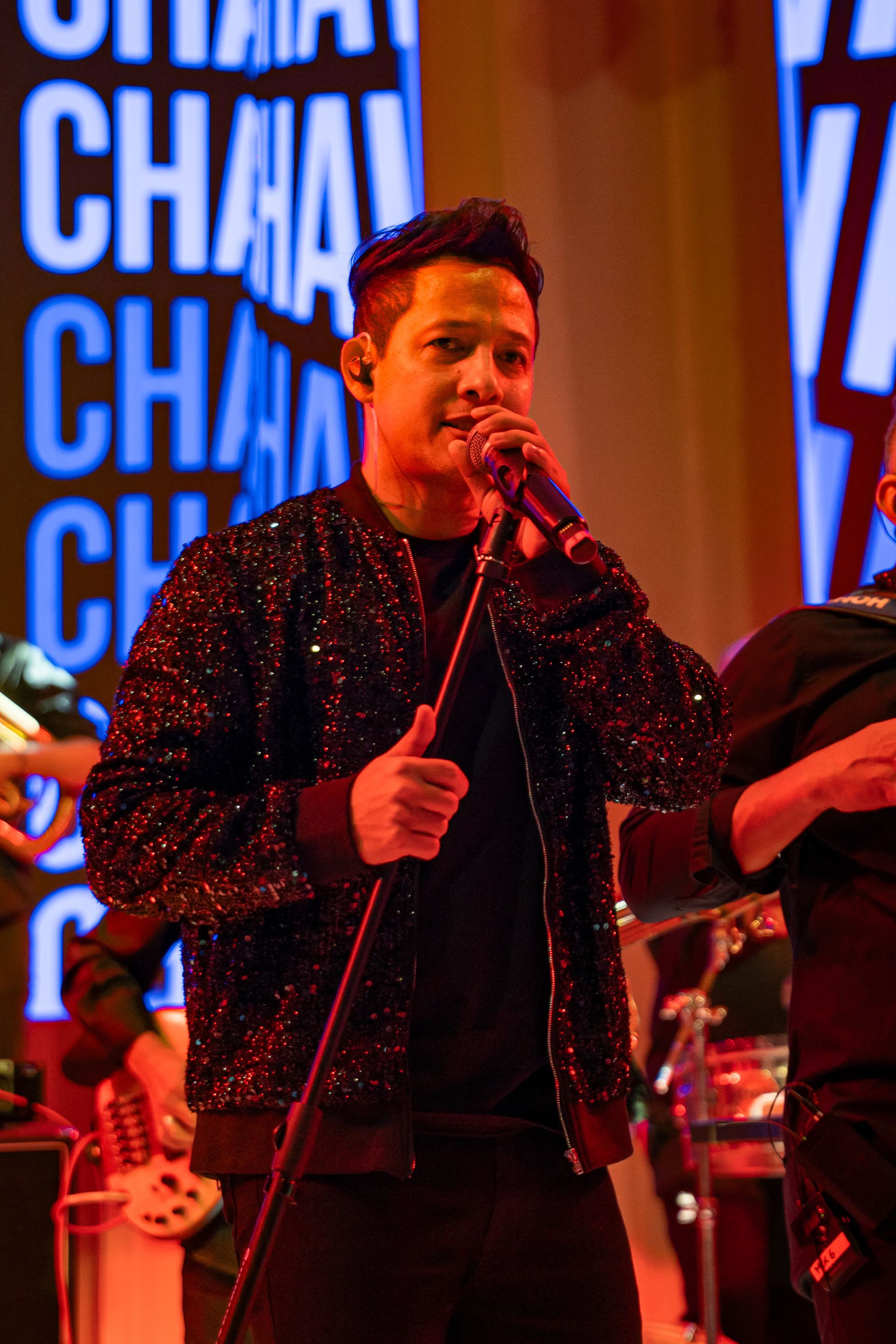 Cantante actuando en un escenario con un micrófono, iluminado en rojo y azul, frente a un gran telón de fondo.