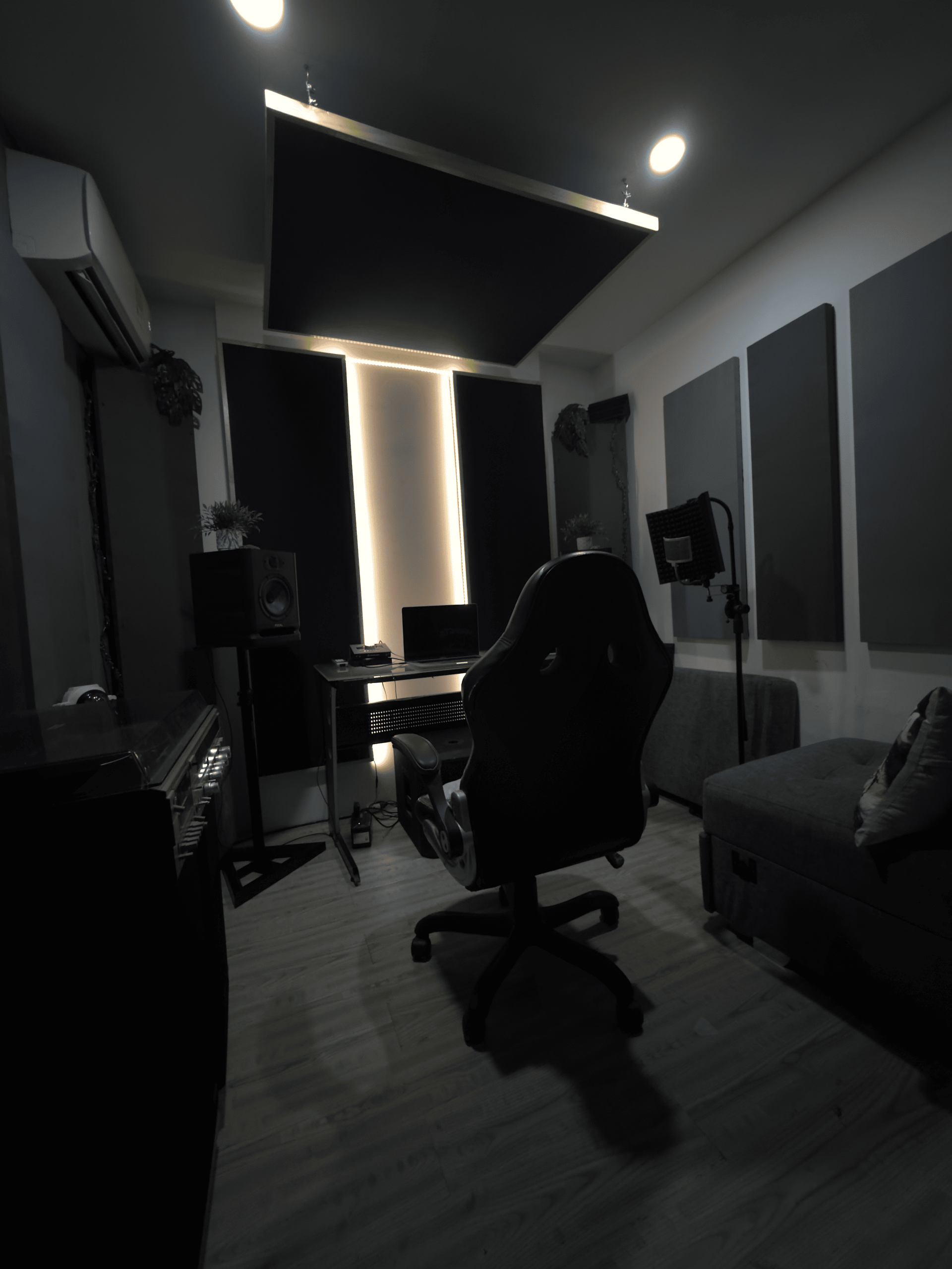 Estudio casero oscuro con un escritorio, una silla, paneles acústicos, un sofá y una ventana iluminada.