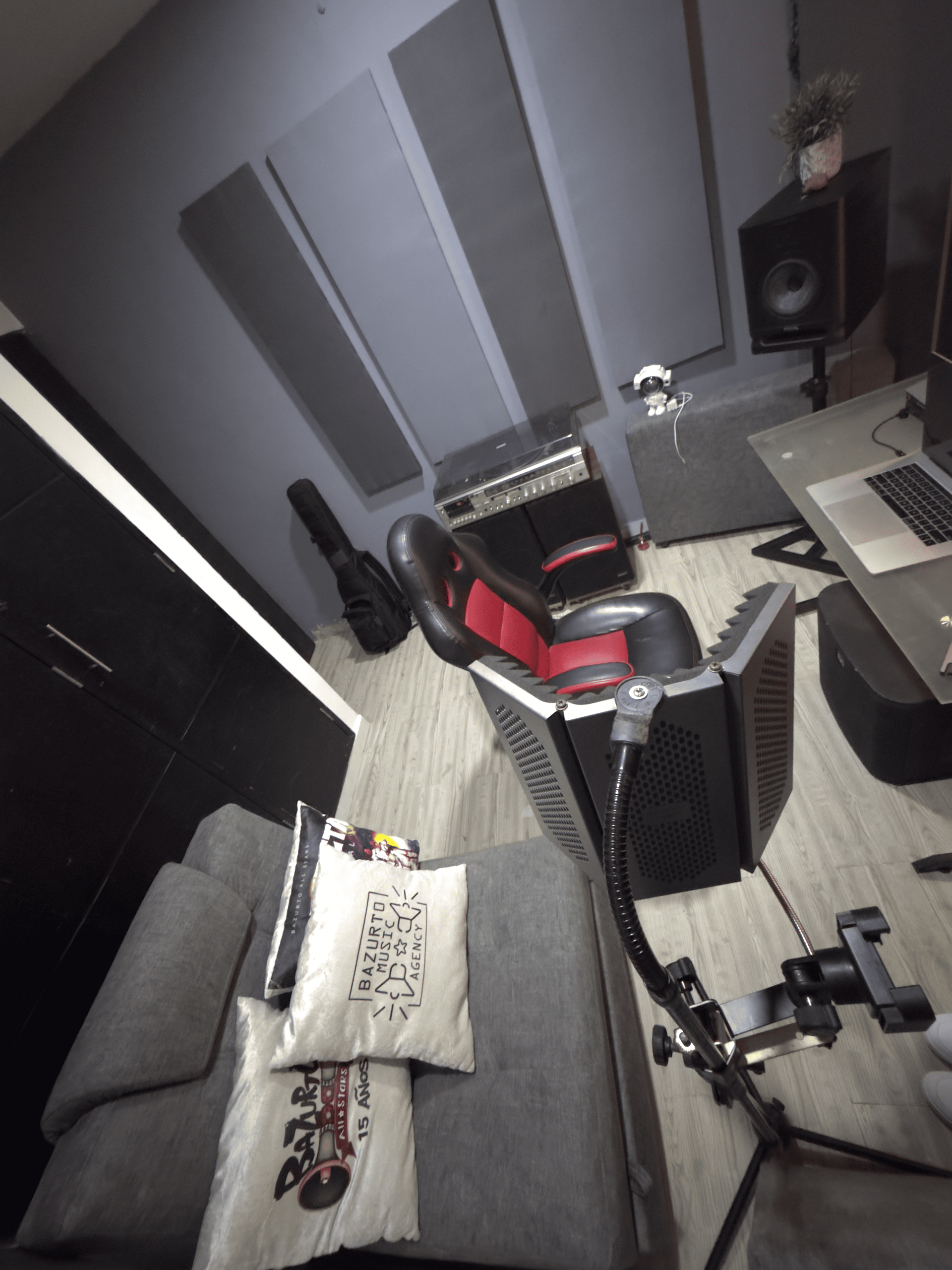 Pequeño estudio con sofá, silla gamer, escritorio, guitarra, altavoz y paneles acústicos en las paredes.