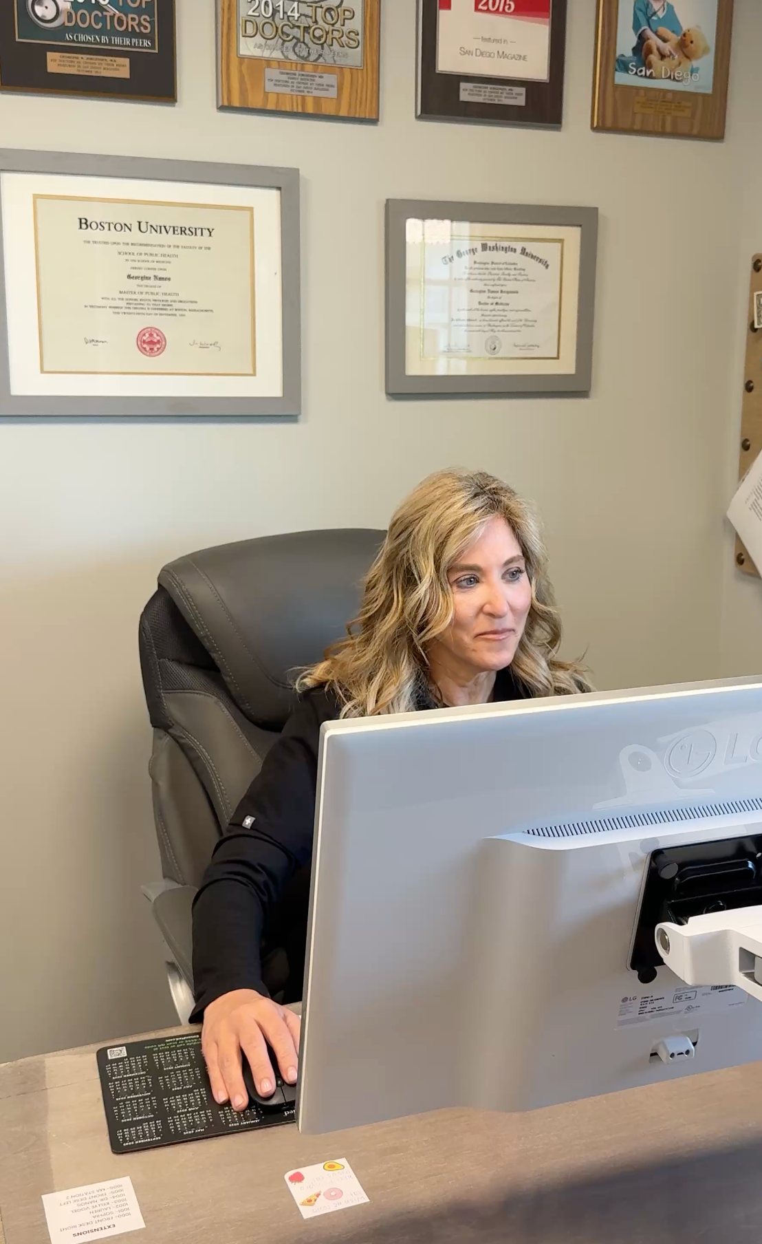 Woman seated at an office desk behind a large monitor, with framed certificates on the wall.