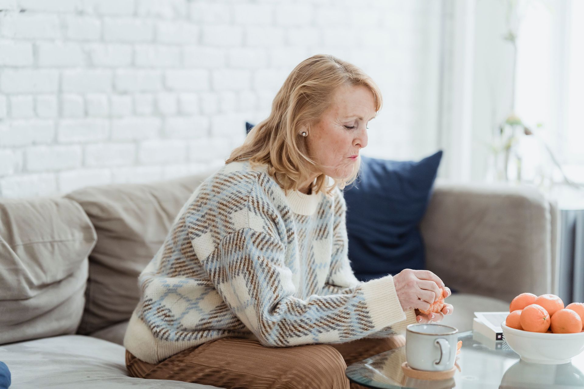 Woman sitting on a couch with a mug and bowl of oranges on a coffee table, looking thoughtful