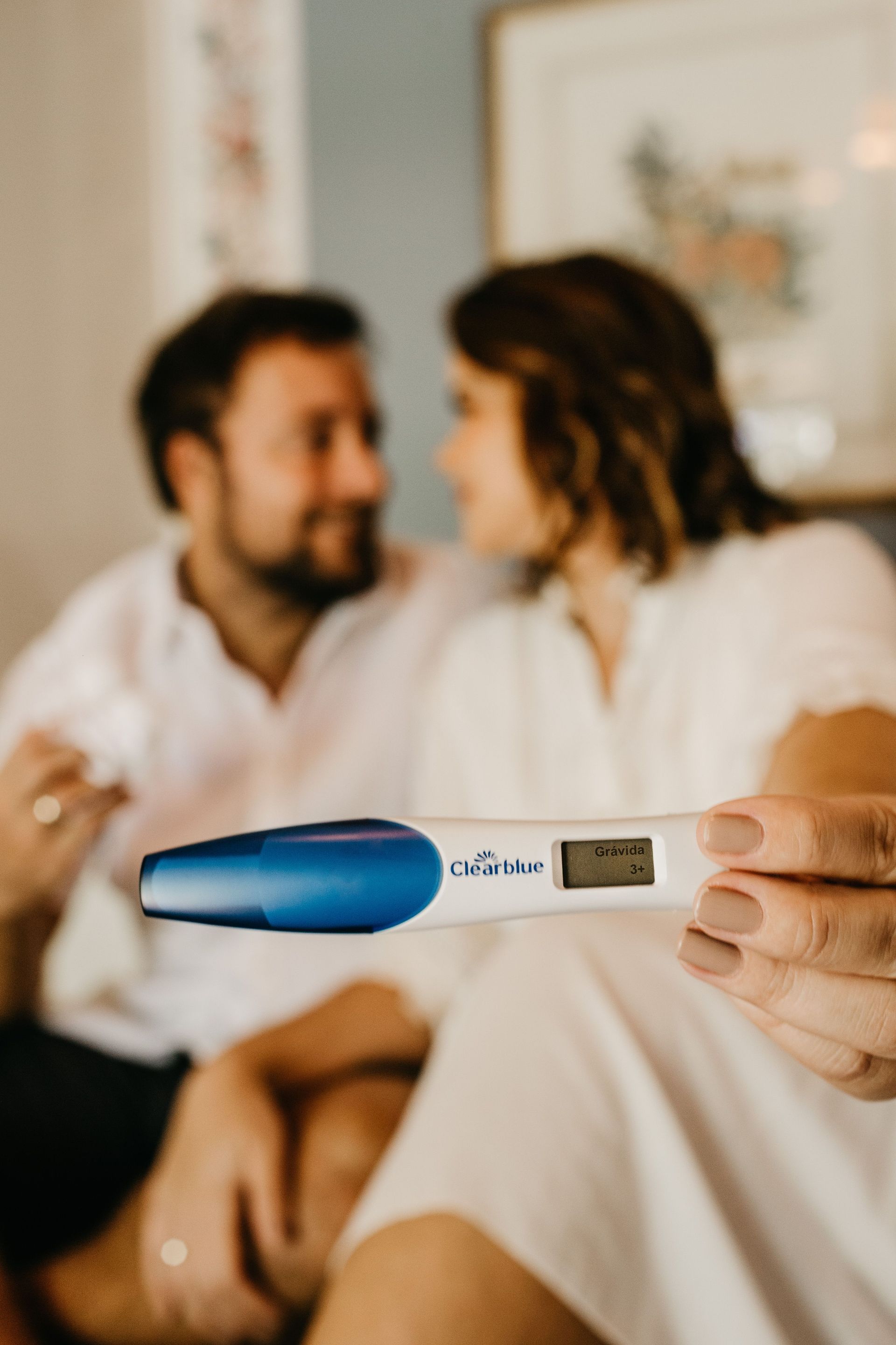 Pregnancy test with blue cap held in foreground, blurred couple sitting together on couch in background