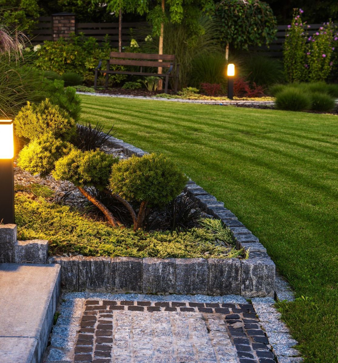 A lush green garden with trees and shrubs and a stone curb.