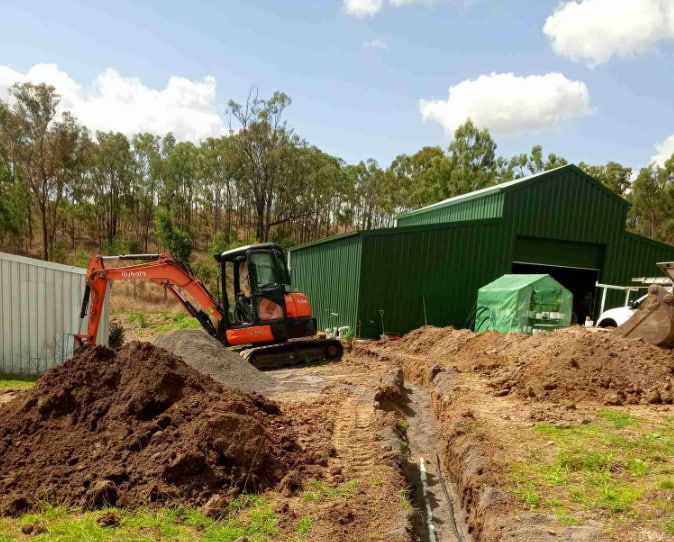 An Excavator is Digging a Hole in Front of a Green Building — Greg Mitchell Plumbing In Nanango, QLD
