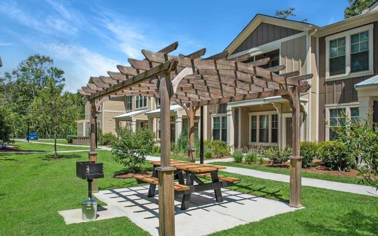Wooden pergola with picnic table and grill in front of a tan building with green lawn.
