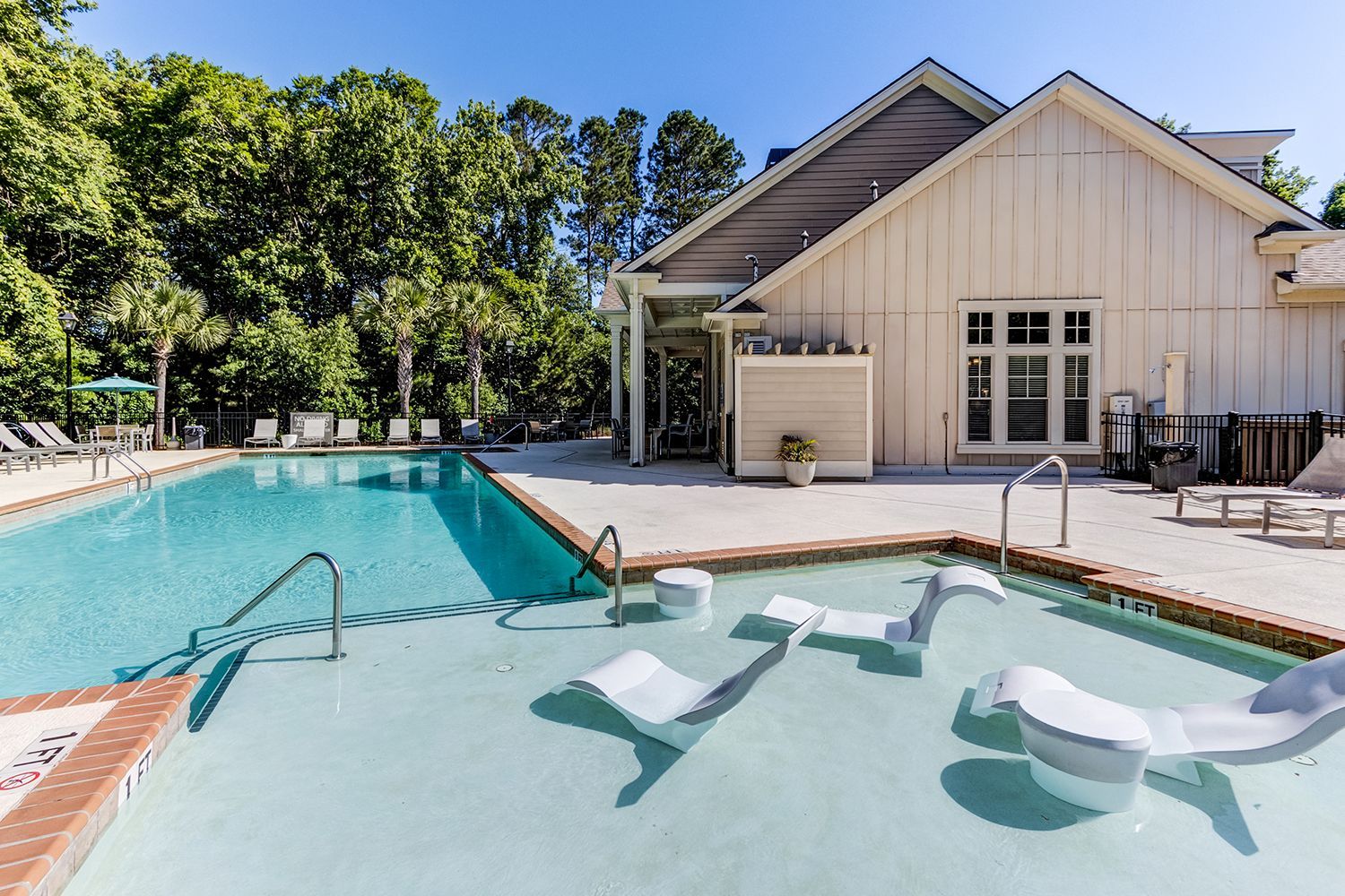 Swimming pool with in-pool loungers, brick border, and a white building in the background on a sunny day at The Preserve at Essex Farms, offers apartments in Charleston, SC.