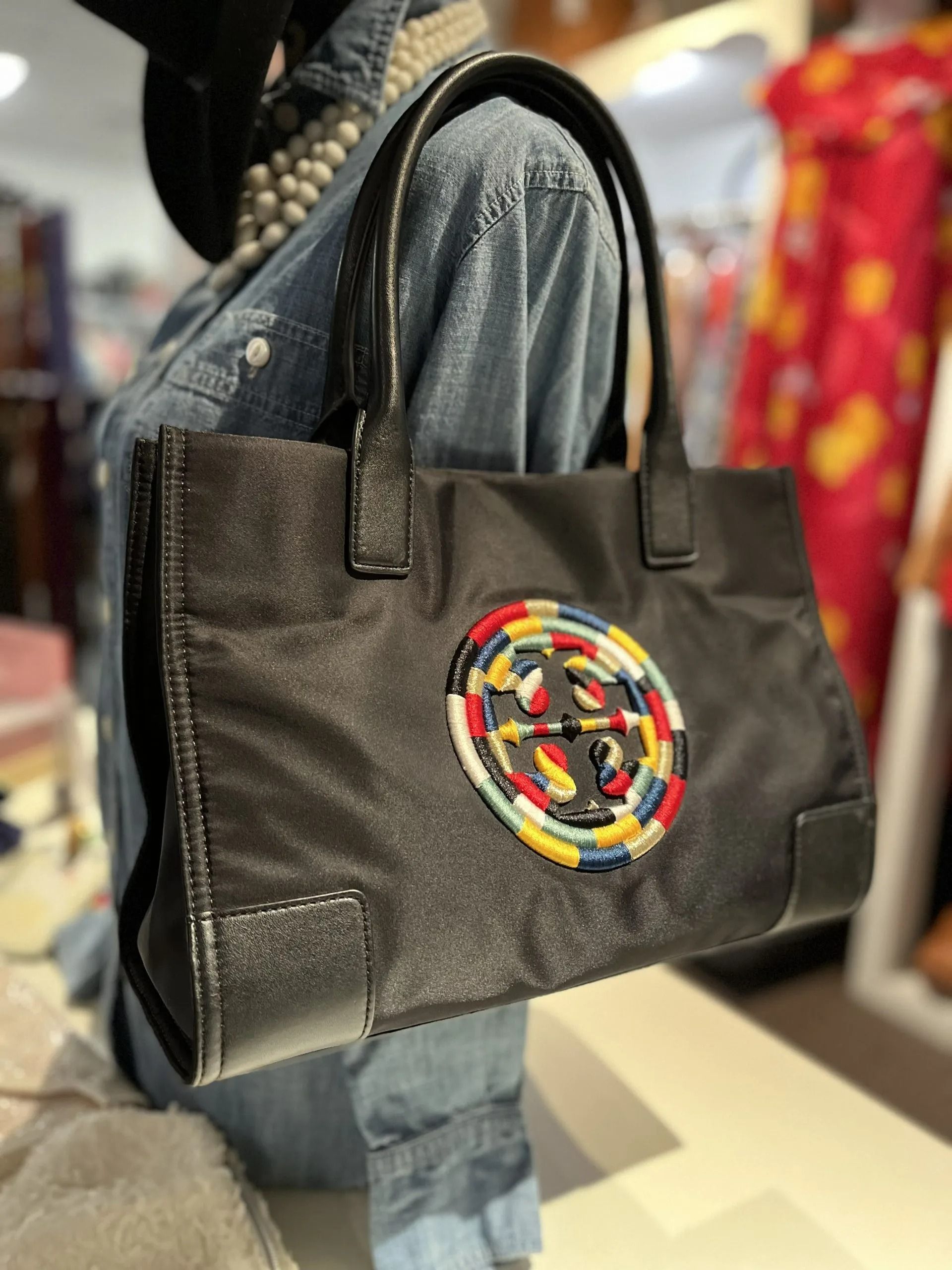 A black nylon tote bag with leather trim and a multicolored circular embroidered logo emblem, displayed on a mannequin.