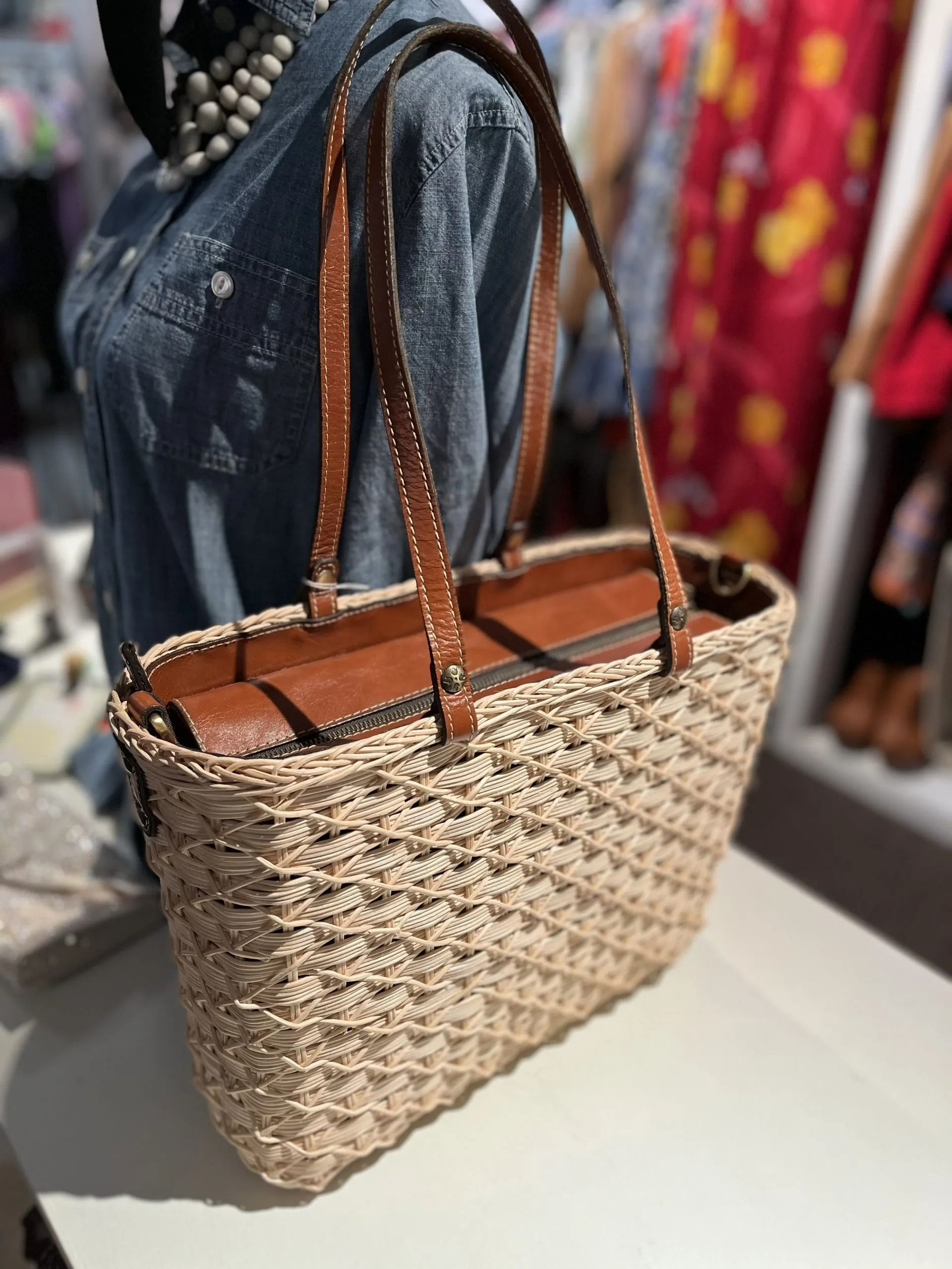 A straw tote bag with brown leather straps and a zipped top, displayed on a counter in a retail clothing store.