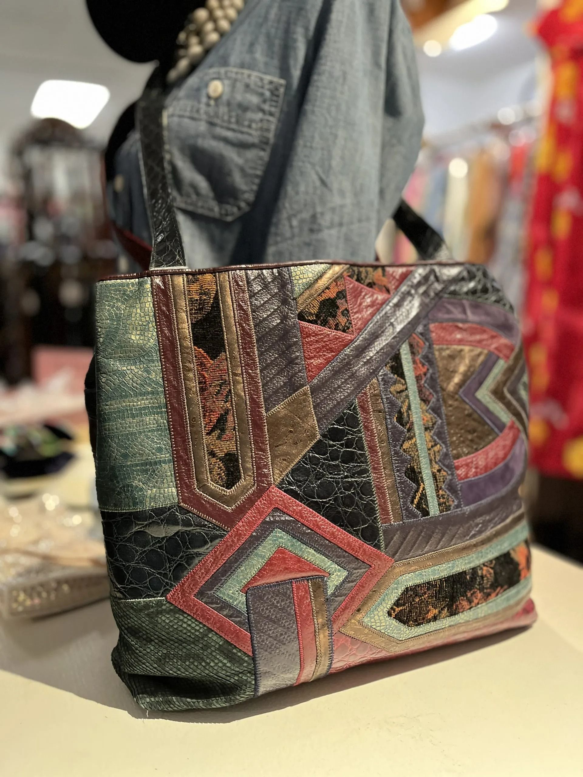 A colorful patchwork-style shoulder bag with intricate, textured patterns in hues of green, red, purple, and gold.