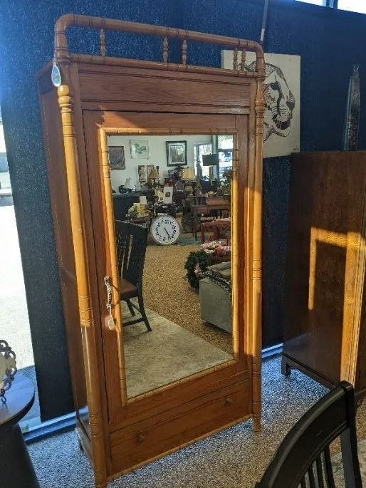 A wooden armoire with a mirror door and a decorative rail on top, standing in a furniture store.