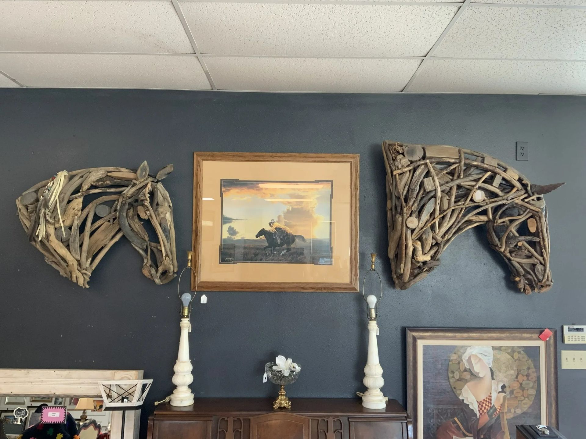 Two driftwood-style horse head sculptures flank a framed western painting on a dark wall above a wooden cabinet.