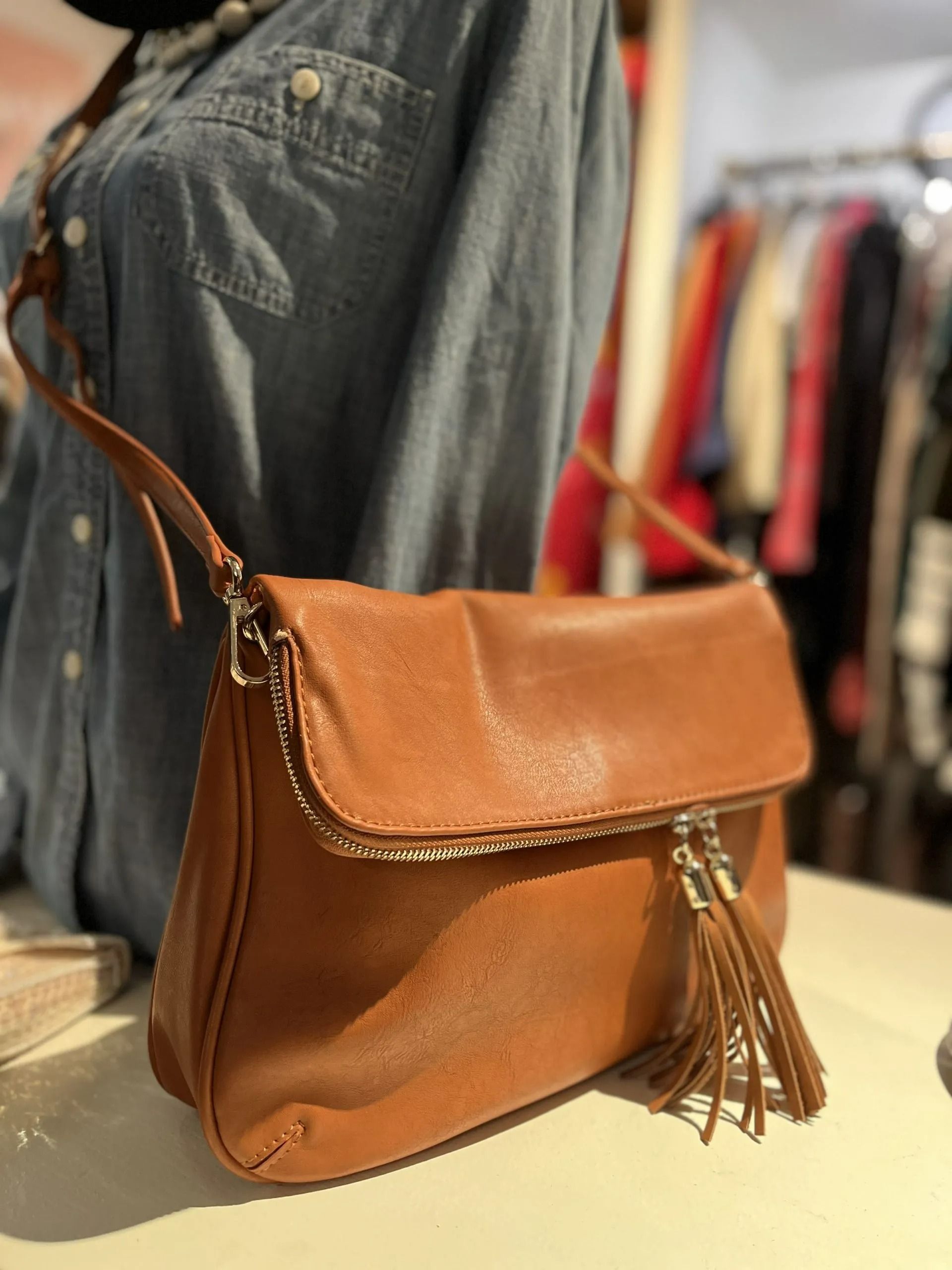 A tan leather crossbody bag with a fold-over flap, decorative stitching, and a dangling fringe tassel.