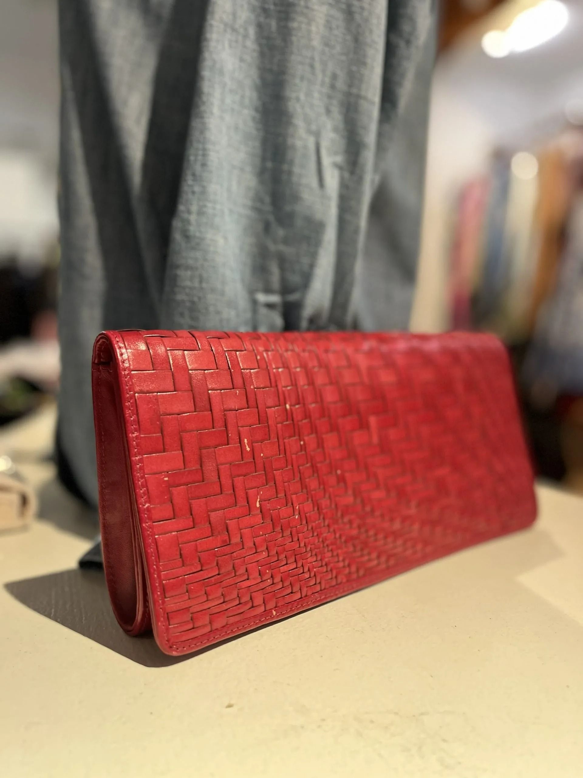 A red, rectangular woven leather clutch purse resting on a light surface with blurred clothing in the background.
