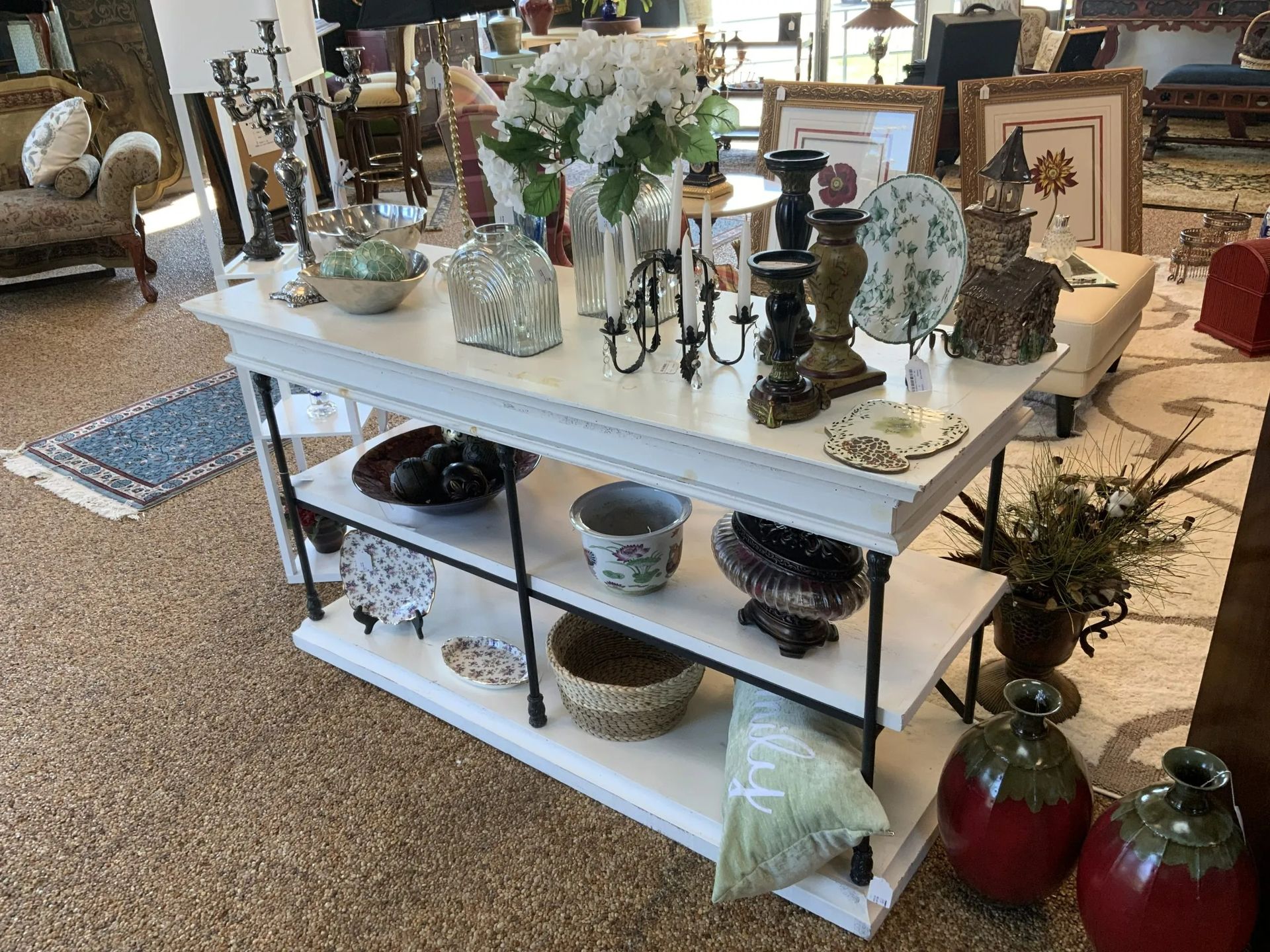 A white, tiered display table in a store featuring decor items like vases, candles, and a bowl of spheres on its shelves.
