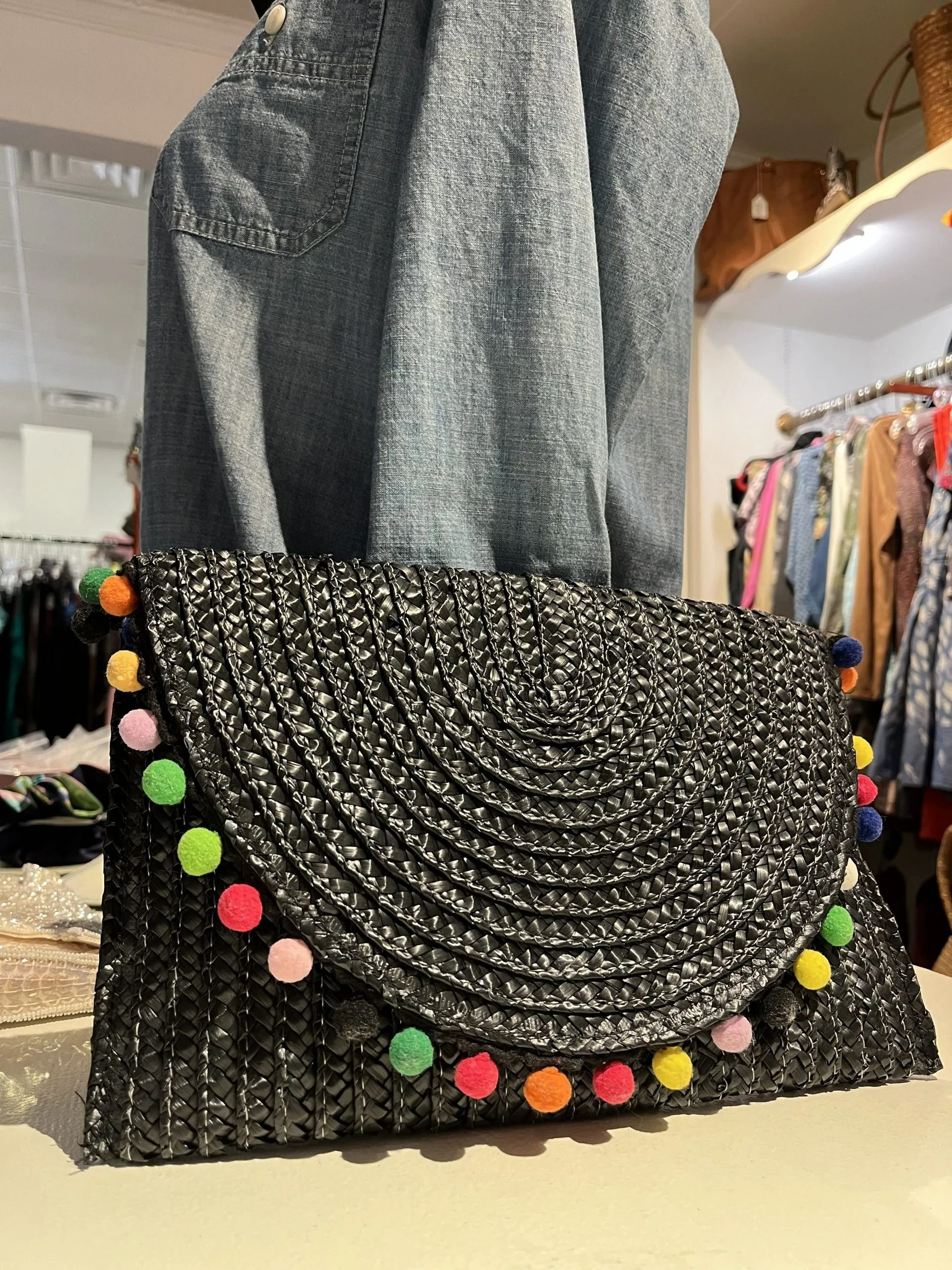 A black, woven straw clutch with a semi-circular flap adorned with colorful pom-poms, displayed in a clothing store.
