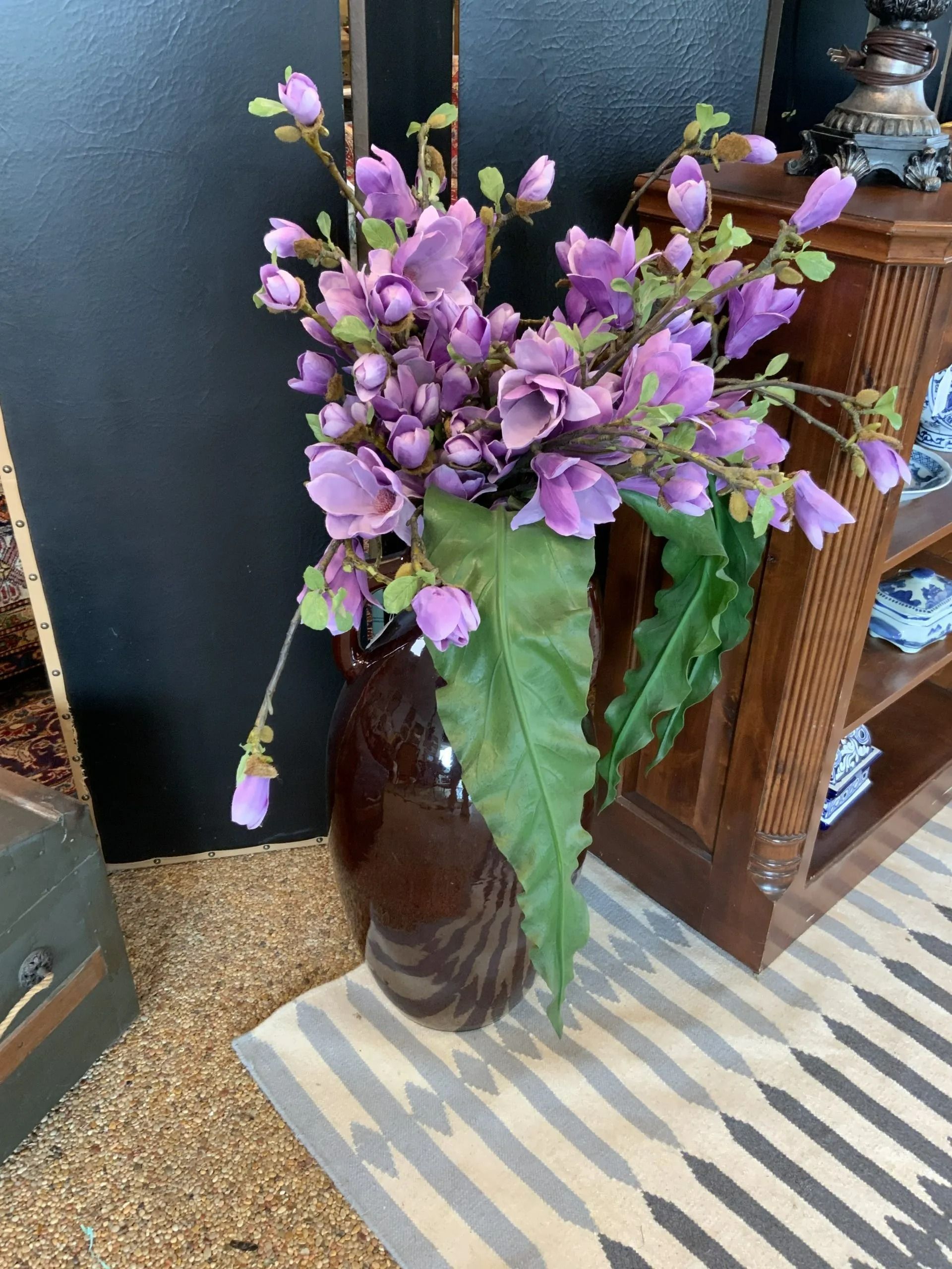 A brown ceramic vase filled with purple artificial flowers and large green leaves, sitting on a patterned rug.
