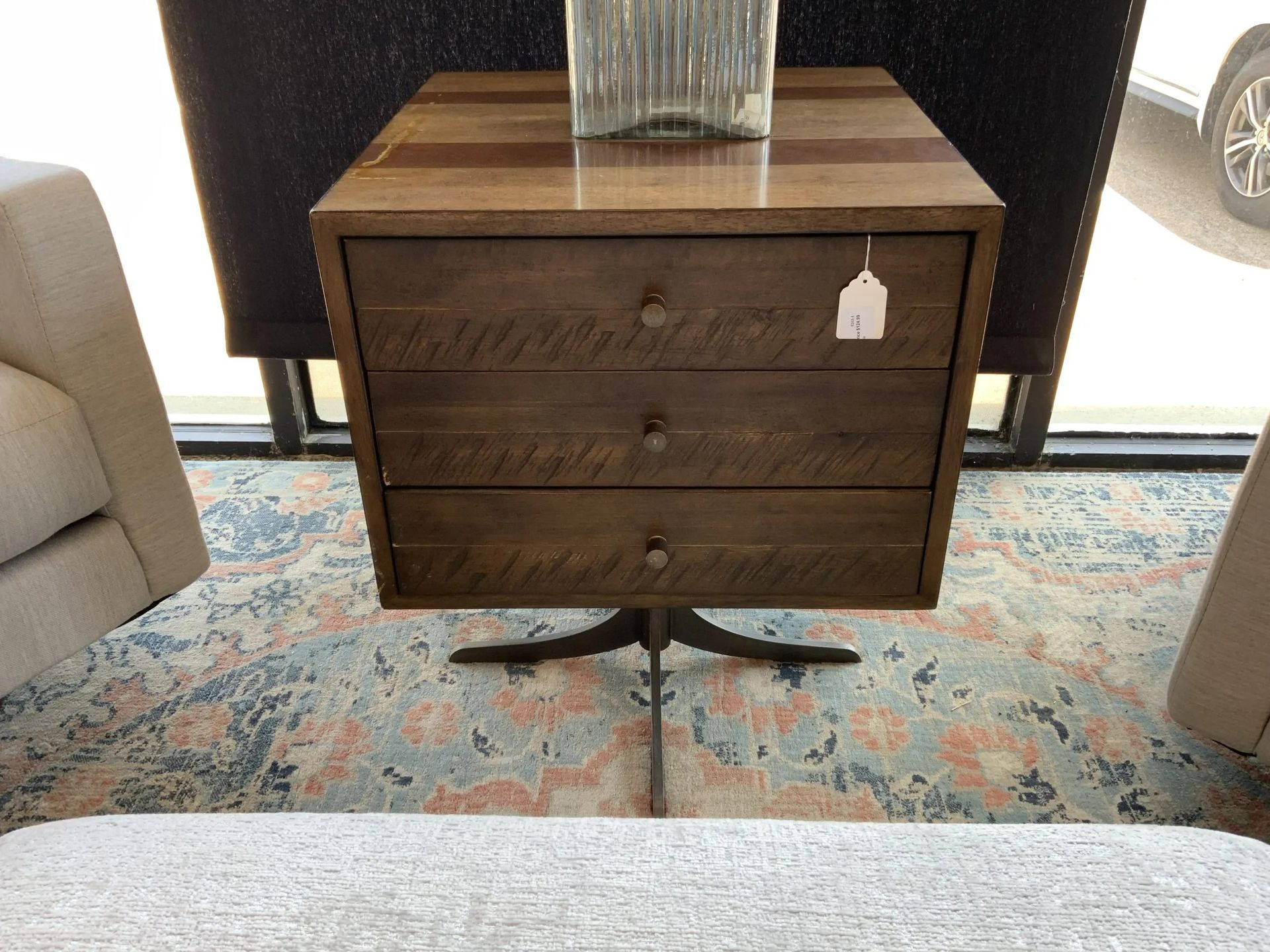 A three-drawer wooden nightstand on a metal pedestal base, placed on a patterned rug in a furniture showroom.
