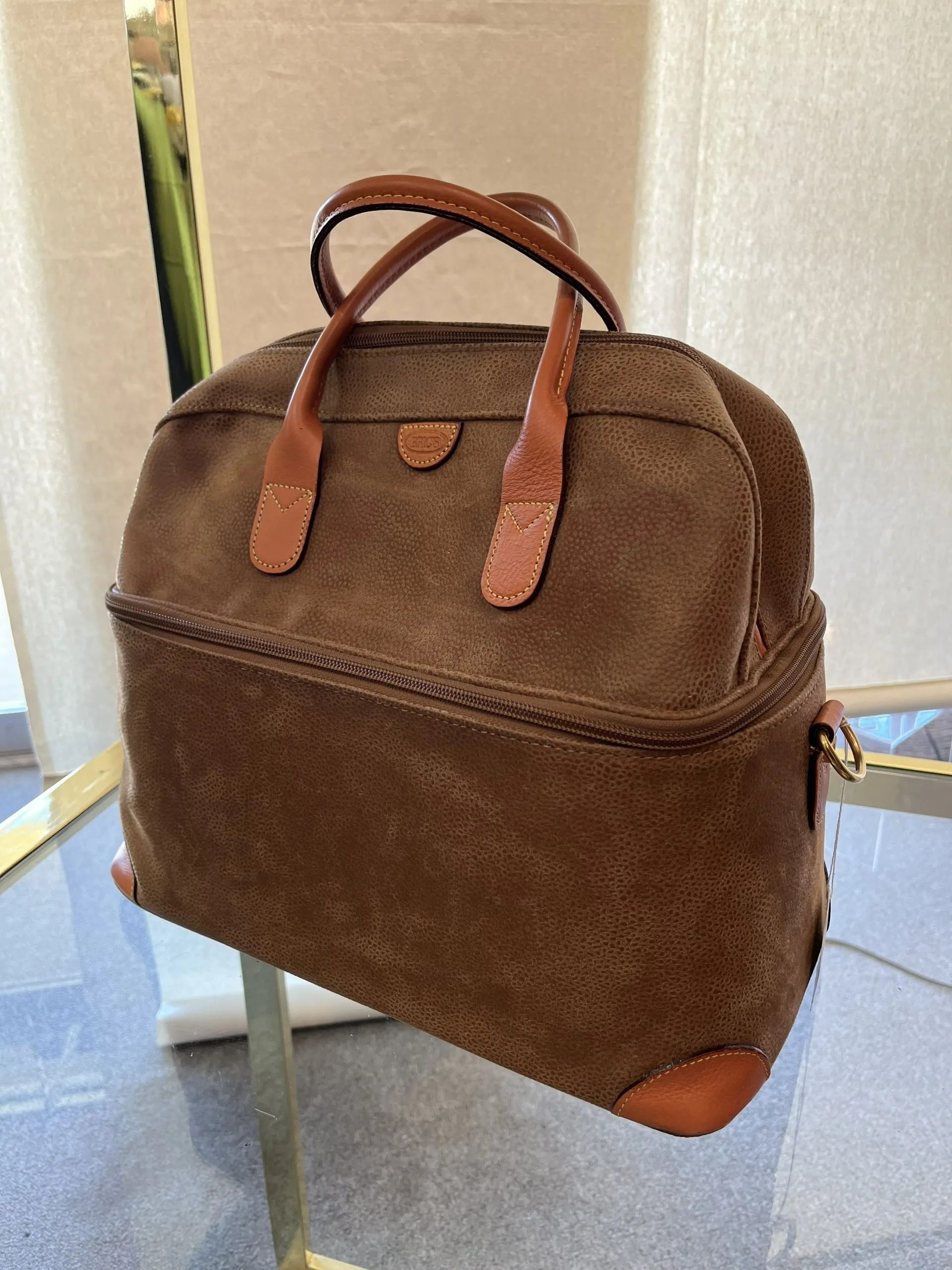 A brown suede handbag with leather handles and accents, resting on a glass display stand.