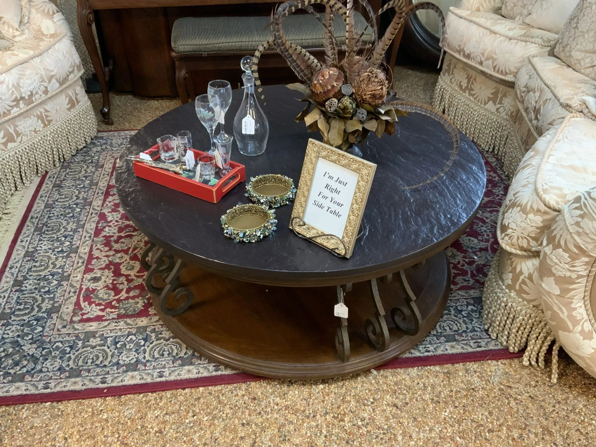 A round, dark wood coffee table with scroll-style legs sits on a patterned rug, decorated with a decanter and small sign.