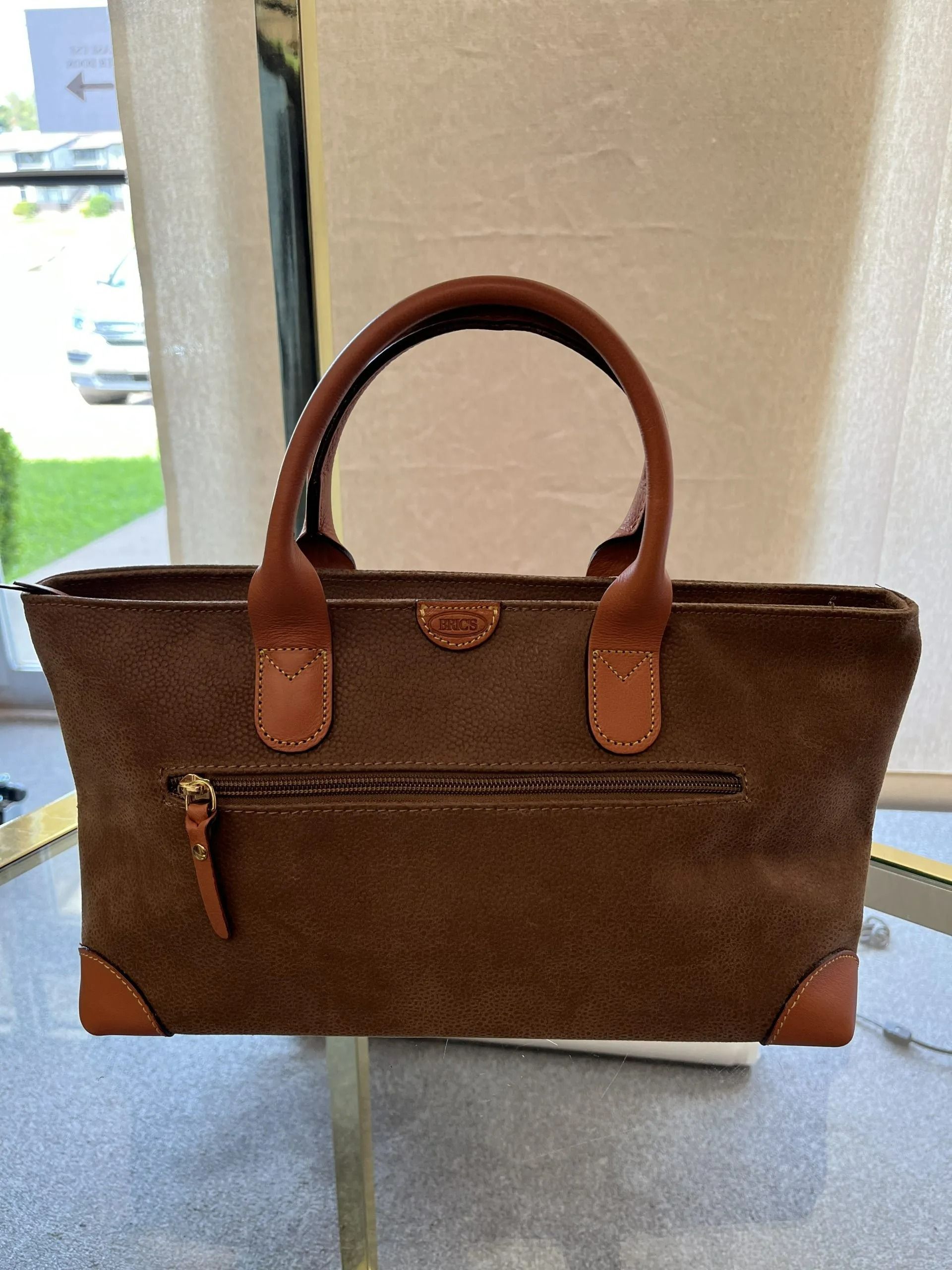 A brown suede handbag with leather handles, corner accents, and a front zippered pocket sitting on a glass surface.