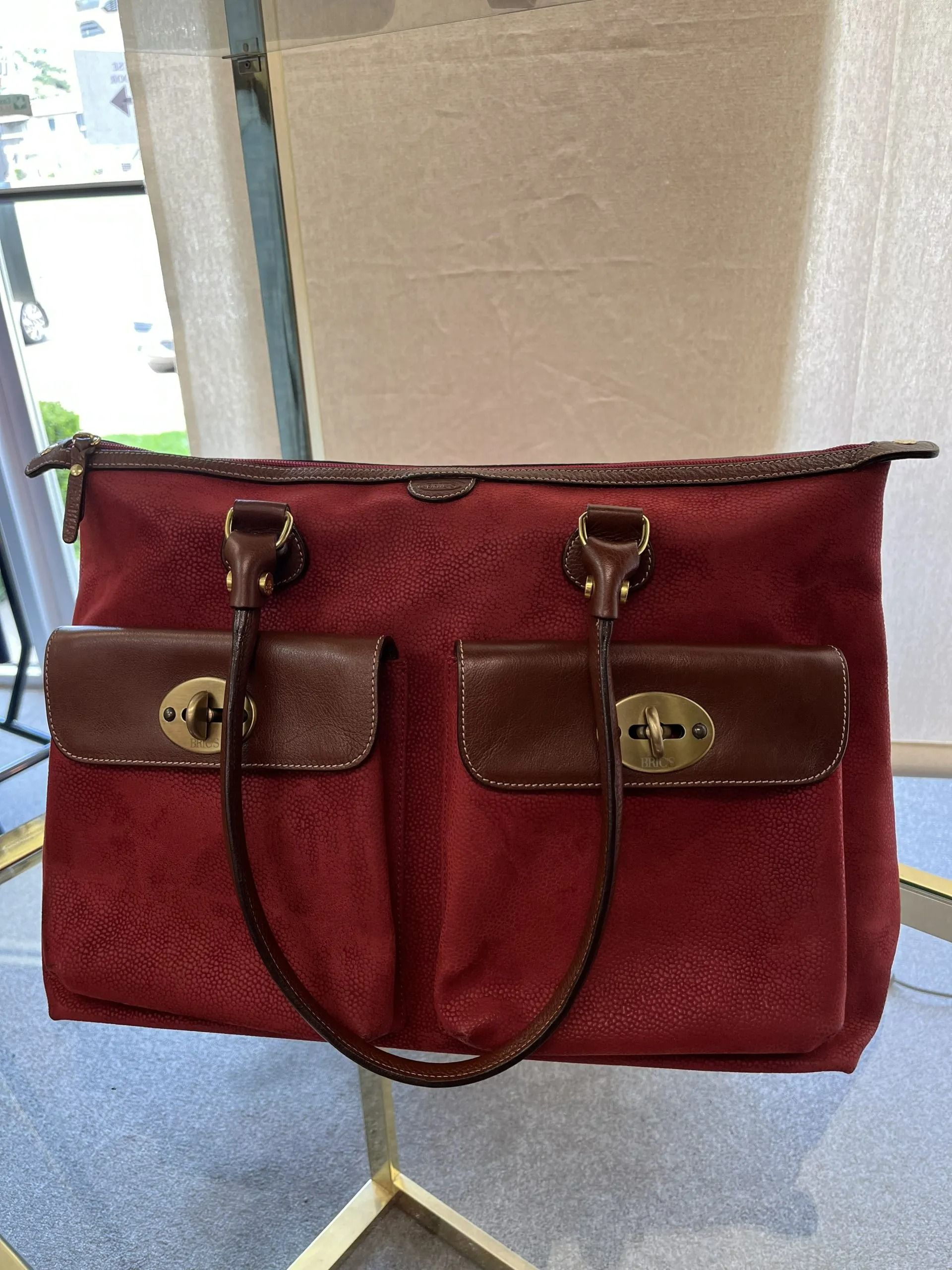 A red, textured tote bag with two brown leather front pockets and brass turn-lock closures on a display stand.