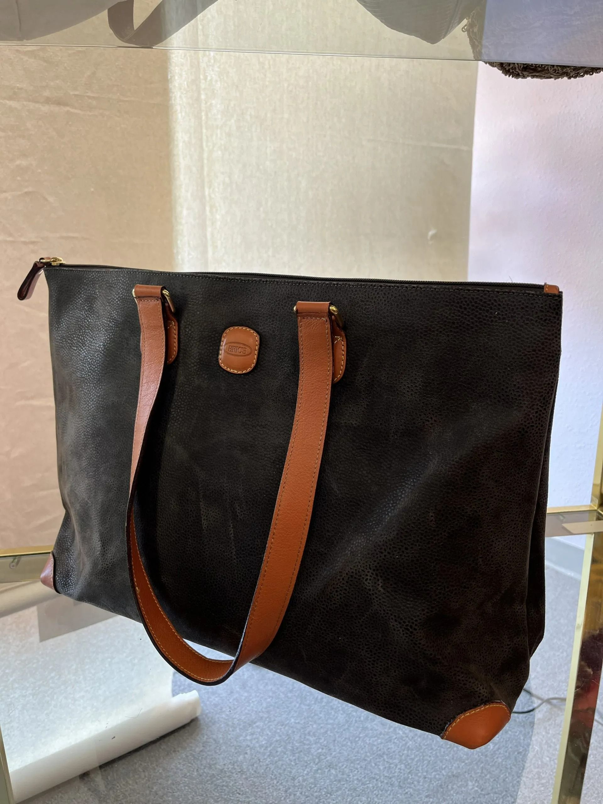 A dark, textured tote bag with tan leather straps and trim, featuring a small circular logo on the center front.