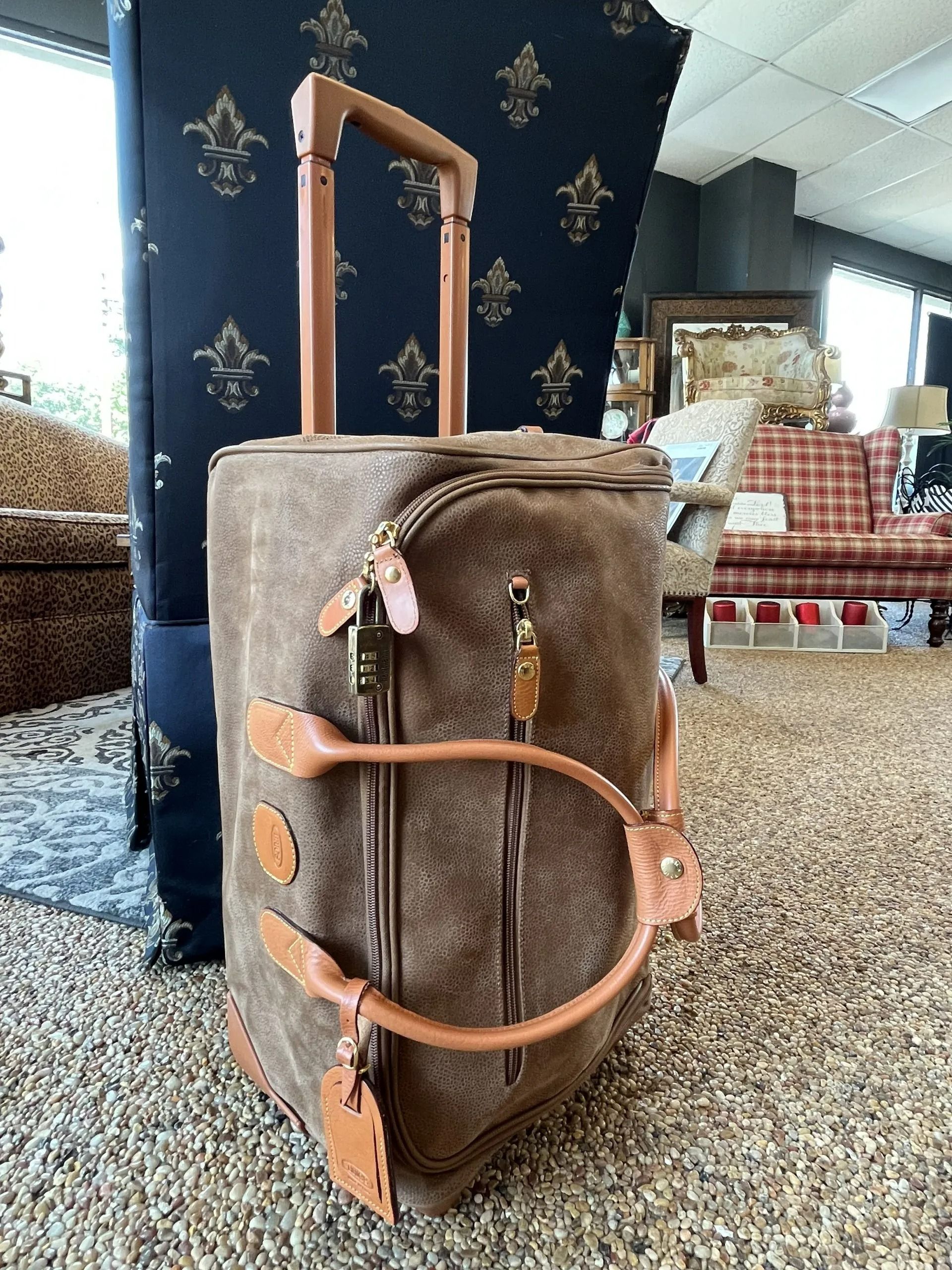 A brown, textured rolling suitcase with tan leather handles and trim, standing on a carpeted floor in a store.